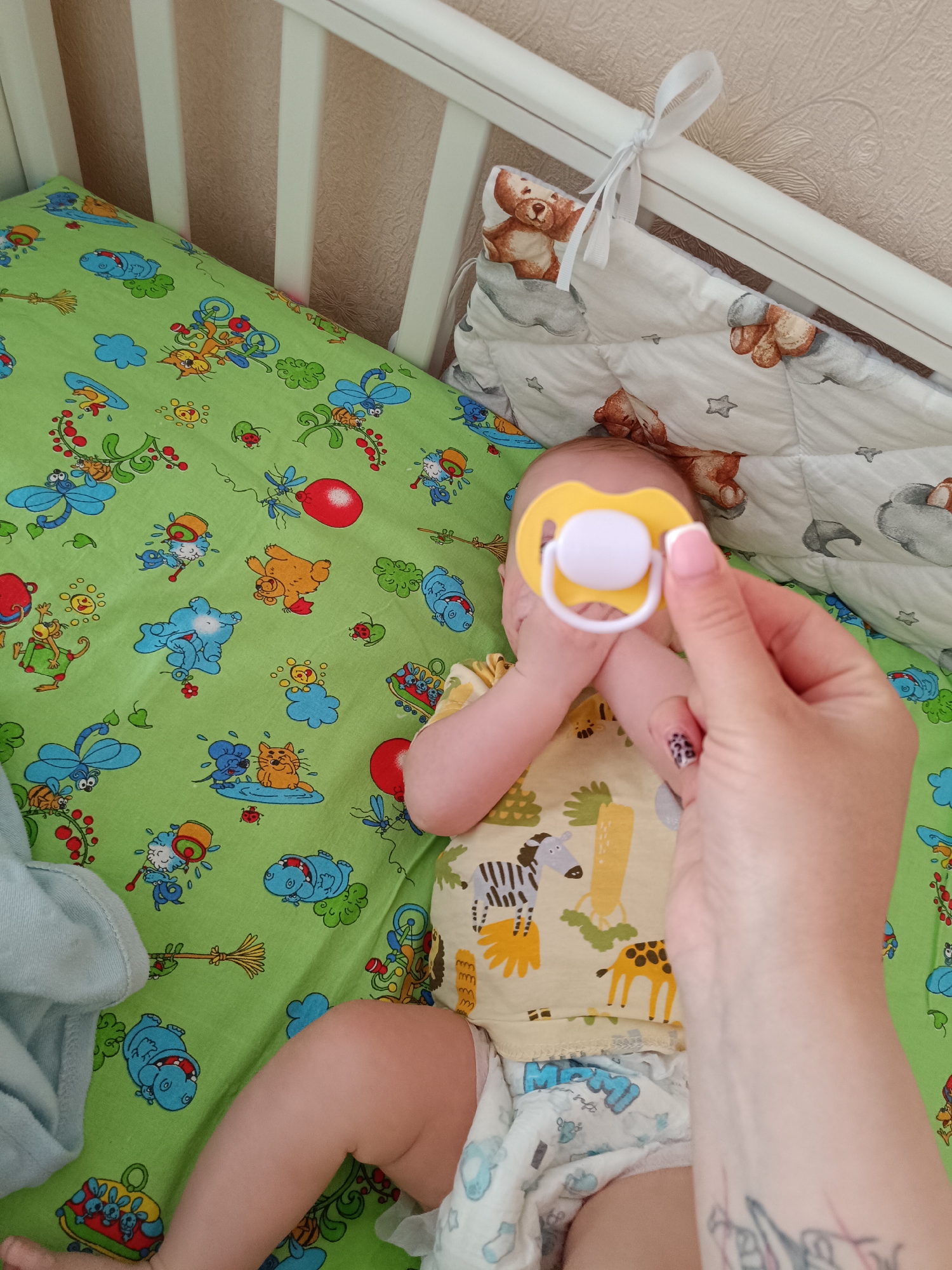 A baby is lying in a crib with a green patterned sheet. The baby is wearing a yellow onesie with animal prints and a diaper. A hand holding a yellow and white pacifier is extended towards the baby. The crib has a white frame and a cushioned bumper with a bear pattern. The crib is located indoors, likely in a home or childcare setting, but there is no confirmation about the city of Tobolsk, Russia.