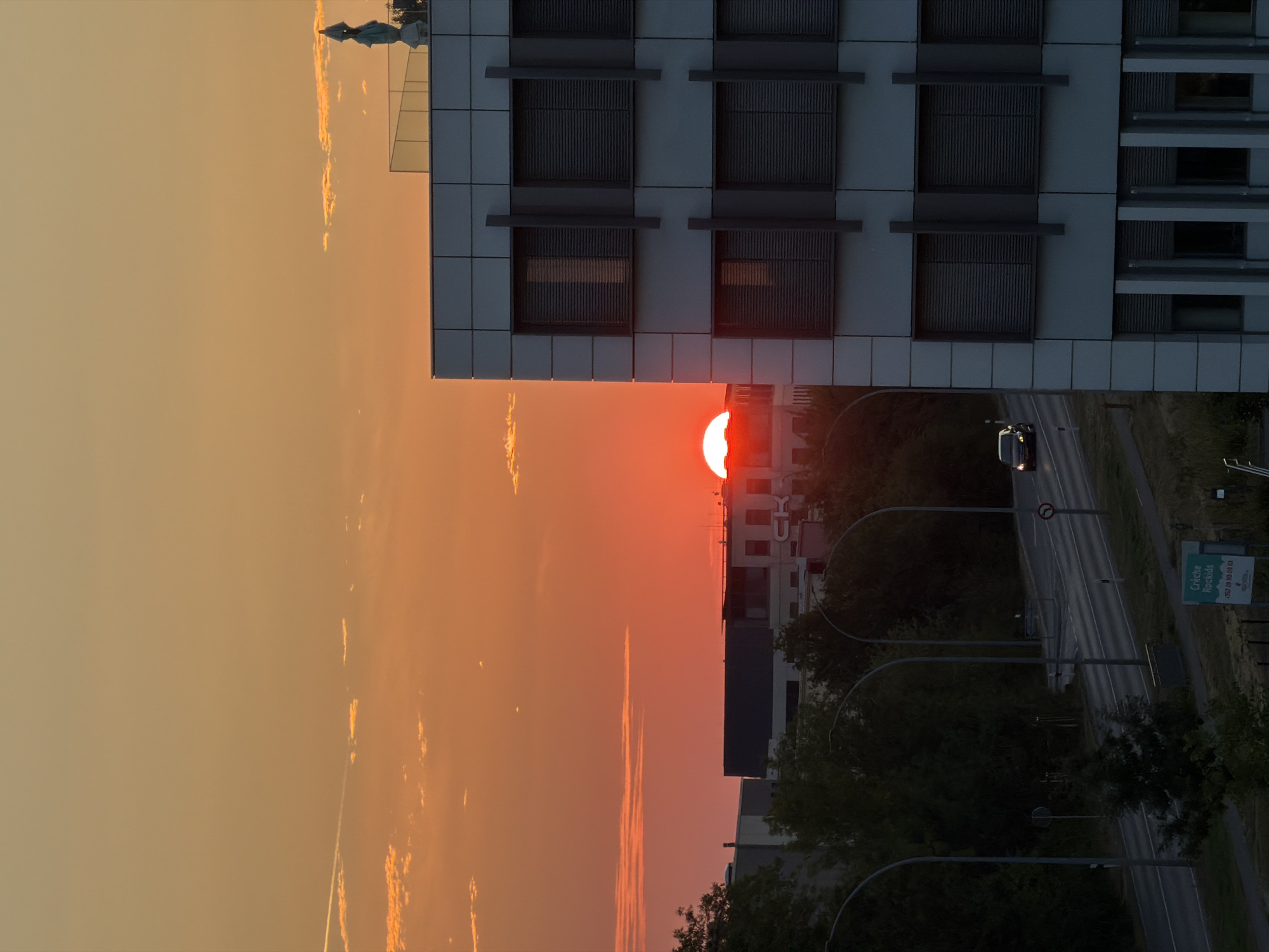The image depicts a sunset over the town of Leudelange, Luxembourg. The sun, a vibrant red orb, is partially obscured by buildings in the distance and a modern office building in the foreground. The sky is a gradient of orange and yellow, with scattered clouds illuminated by the setting sun. The office building features a facade of white panels and dark-framed windows with blinds. A small statue of a figure in blue is visible on the roof of the building. In the mid-ground, a road with streetlights runs past trees and other buildings. A car is visible driving on the road. A sign in the lower right corner reads "Crèche Rockids". The overall scene is one of an urban environment at dusk, with the natural beauty of the sunset contrasting with the modern architecture.