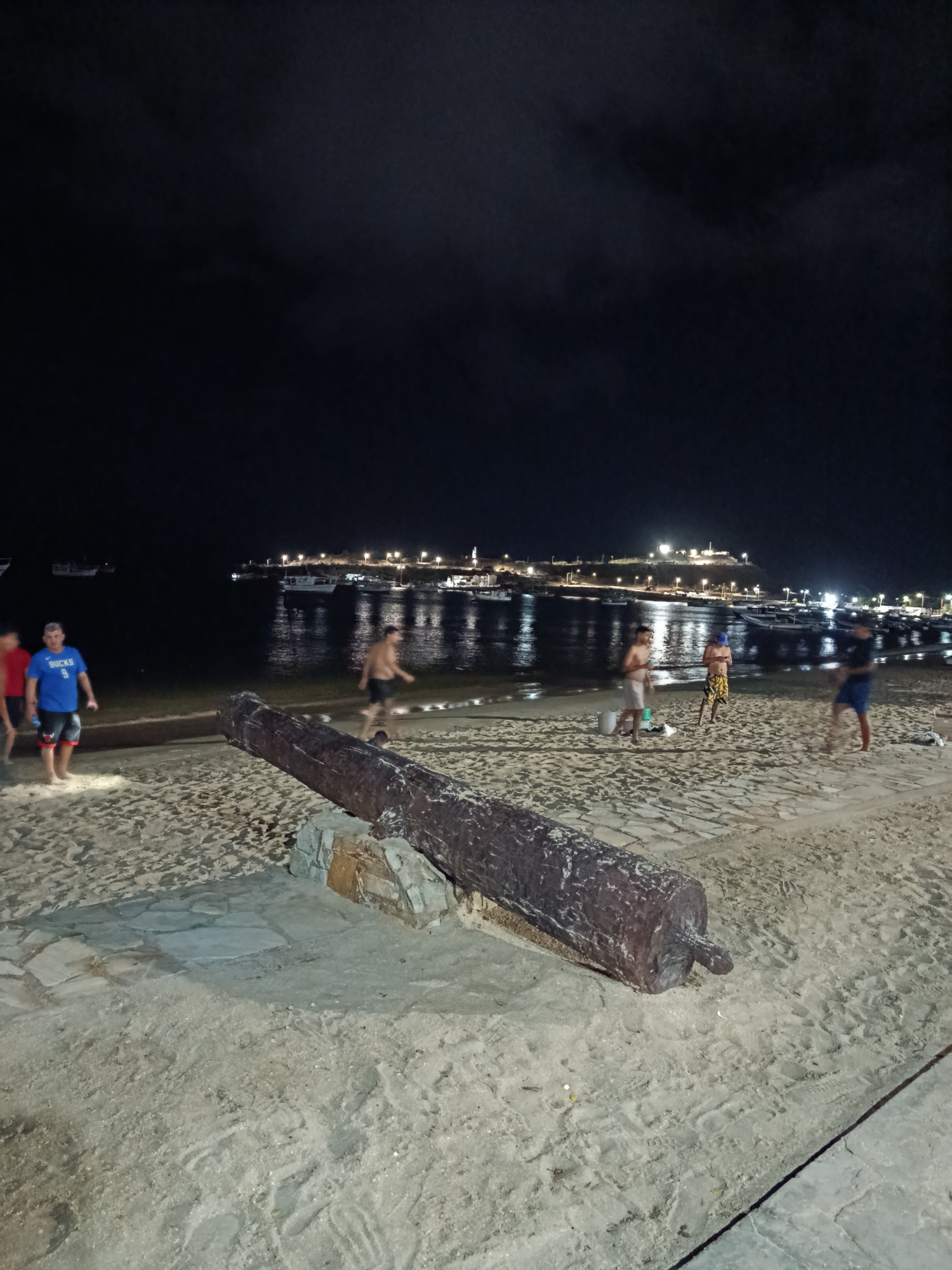 The image depicts a nighttime scene on a sandy beach in Juan Griego, Venezuela, with a body of water in the background. In the foreground, a large, weathered cannon rests on the sand, supported by stones. Several people are visible on the beach and near the water's edge. On the left, two men stand on the sand, one wearing a blue t-shirt and shorts. Further back, near the water, several individuals are engaged in various activities; some appear to be playing or walking. In the middle ground, the calm water reflects the lights of boats and buildings lining the distant shore. The shoreline is dotted with numerous small boats, and a town or settlement with illuminated buildings and structures, possibly including a lighthouse or prominent tower, is visible on a hill overlooking the bay. The sky is dark and cloudy, typical of nighttime. The overall atmosphere is relaxed, with people enjoying the beachfront at night.