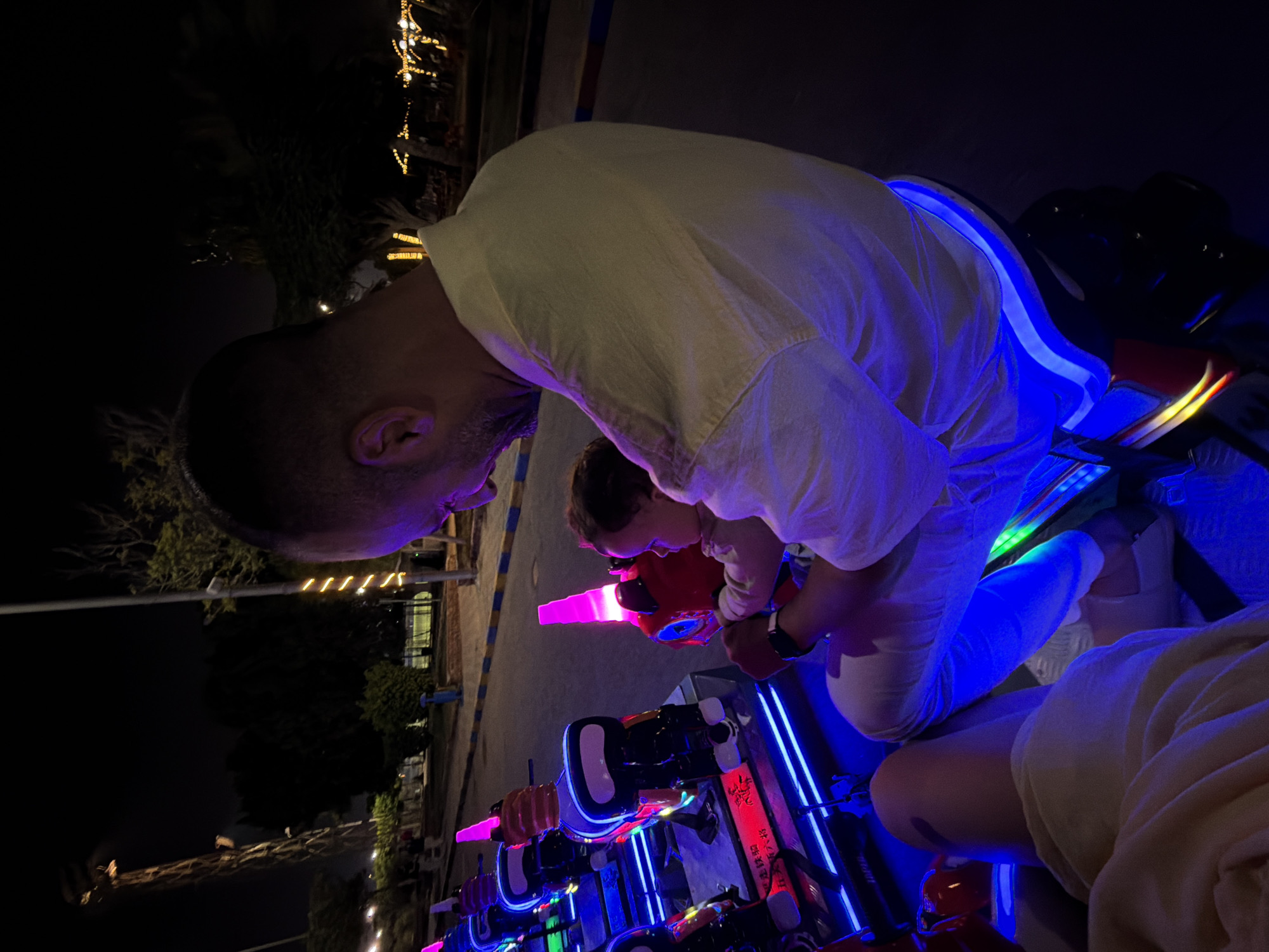 In the city of 6th of October, Egypt, a man and a baby are seated on a ride at night, illuminated by bright neon lights. The man, with a shaved head and a beard, is wearing a light-colored shirt and white pants, and he is holding the baby, who is facing away from the camera and looking at the ride. The baby is wearing a light-colored outfit. The ride consists of several colorful, vehicle-shaped seats, each with bright pink, blue, and red lights. In the background, trees, streetlights, and a tall structure, possibly a Ferris wheel or a tower, are visible. The scene appears to be at an amusement park or a recreational area.