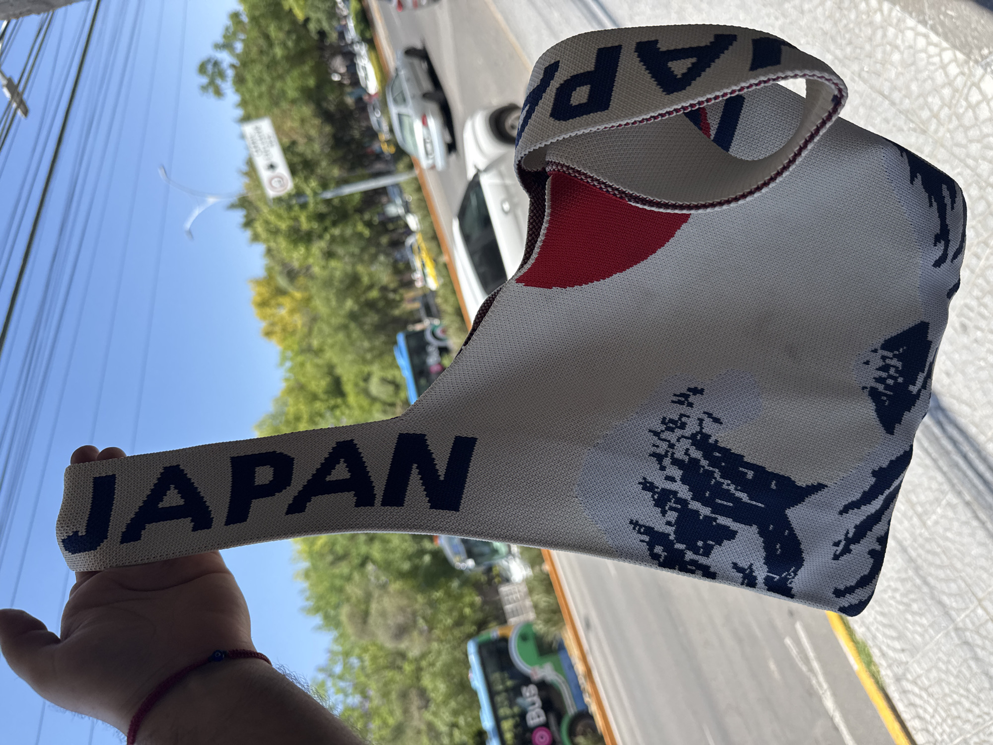 A low-angle, eye-level, medium shot shows a person holding a "Japan" themed cloth bag in the city of Querétaro, Mexico. The bag has a white background with blue "JAPAN" lettering on its handle and around the rim of the bag's opening. There is a red circular design on the bag, and a dark blue stylized pattern on its lower right side. The person's hand, wearing a red bracelet with a blue bead, is visible holding the bag's handle against a partly cloudy blue sky. Overhead power lines are visible in the upper left of the frame.
In the background, slightly out of focus, a street with cars and buses is visible. Green trees line the street. A street sign is mounted on a pole on the median between the lanes of traffic. The bag appears to be held over the side of a building, looking out onto the street.