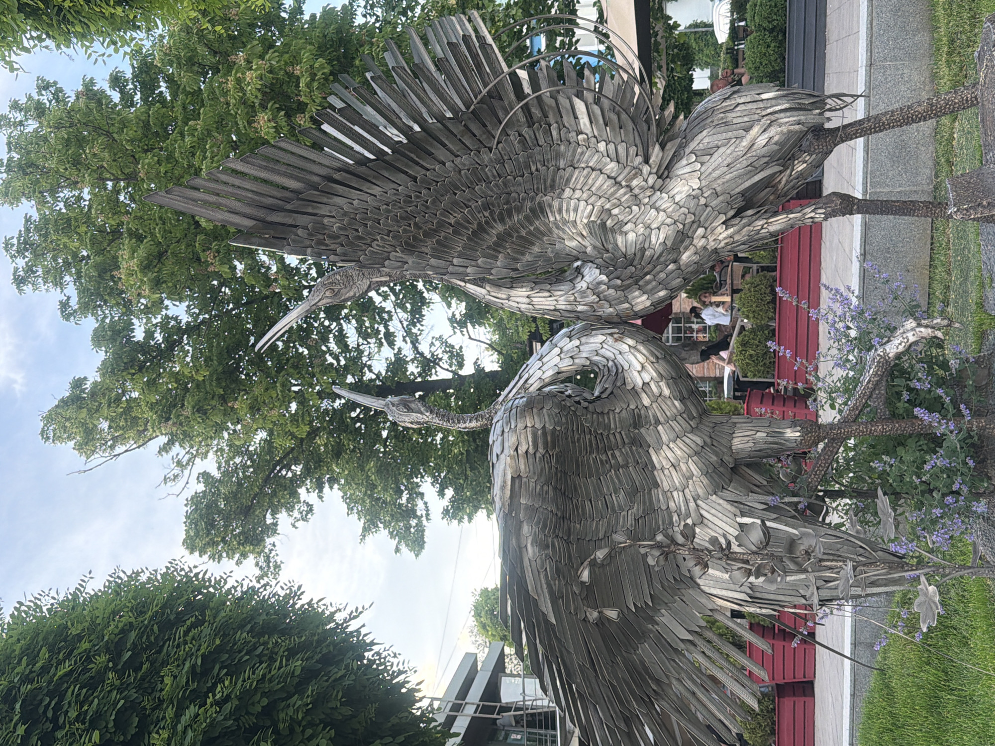 The image shows two silver crane sculptures in Rostov-on-Don, Russia. The cranes stand on a grassy area adjacent to a sidewalk, with a red slatted fence in the background. The sculptures are highly detailed, with individual metal pieces forming the feathers and body structure. Trees and greenery are visible in the background, partially obscuring buildings. A few people are seated on a bench further back in the scene. Purple flowers grow near the base of the sculptures.