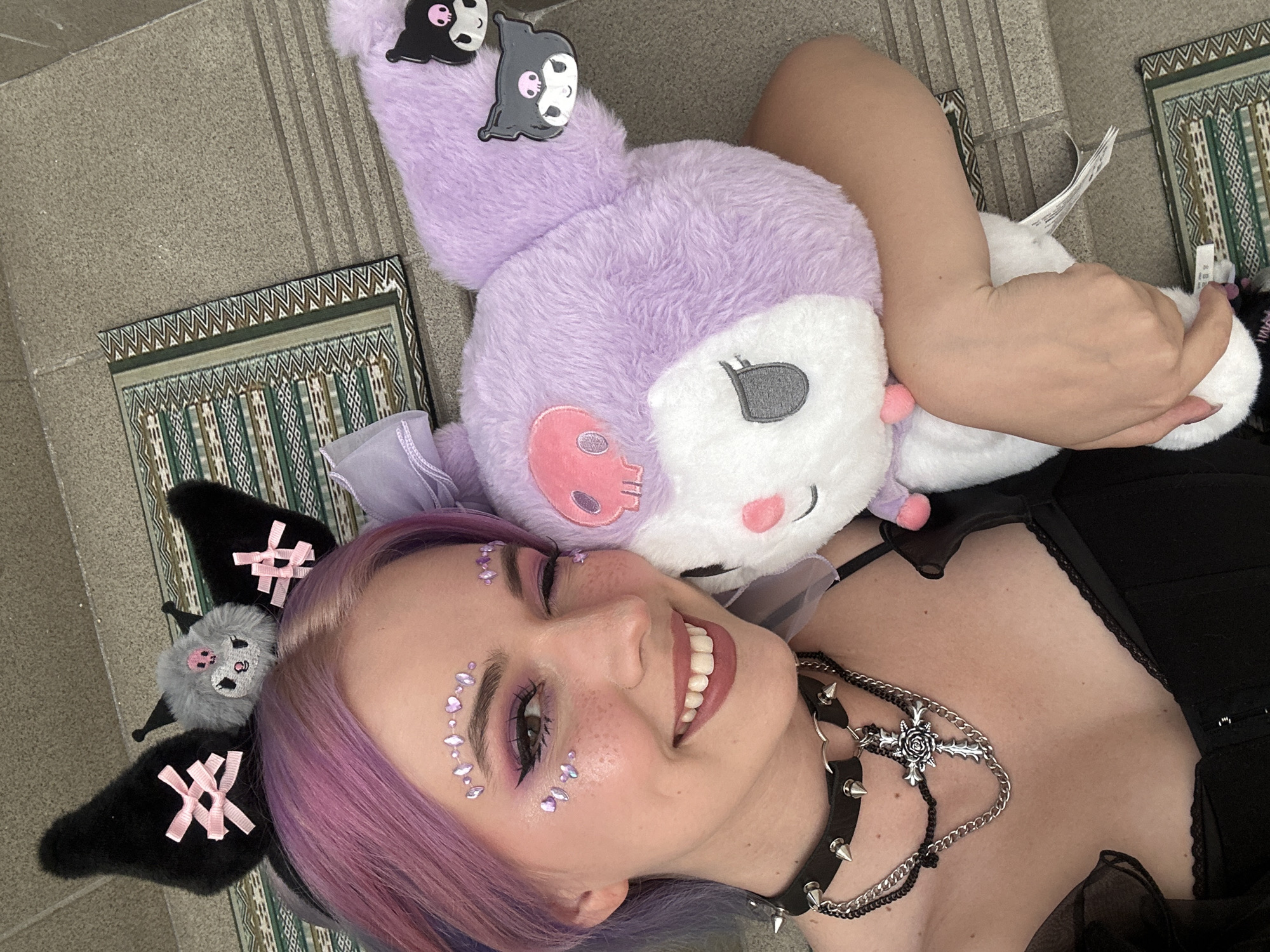 A person with fair skin and purple hair is lying on a tile floor, smiling. The person is wearing black clothing and a spiked choker with a cross necklace. They have purple eye makeup and lavender-colored adhesive gems on their face. They are wearing a black headband with bat wings and pink bows. To the left of the person is a small rectangular rug with a geometric pattern. A large plush toy, lavender in color and resembling a character with black patches, rests partially on the person's head and arm. A white tag is visible on the plush toy. The tile floor is gray and appears to be outdoors. The photograph does not contain sufficient information to determine whether it was taken in the City of Zagreb, Croatia.