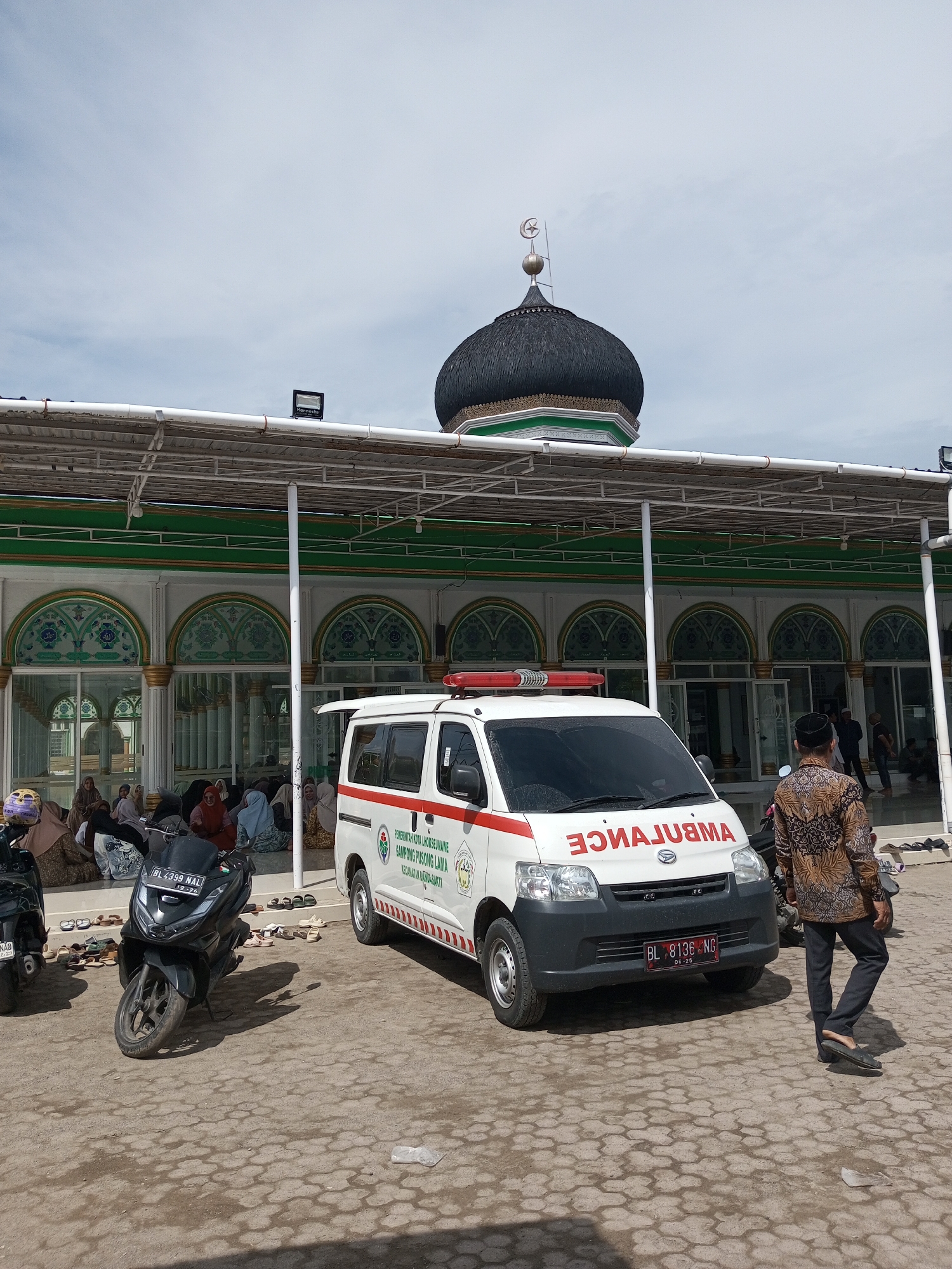 The image shows a mosque in the Gampong Pusong Lama area of the Kota Lhokseumawe region in Indonesia. An ambulance is parked in front of the mosque, and a person wearing traditional Indonesian batik is walking toward the building. Several women are seated inside the mosque. A motorbike is parked nearby. The mosque has a dome topped with a crescent moon and star.
