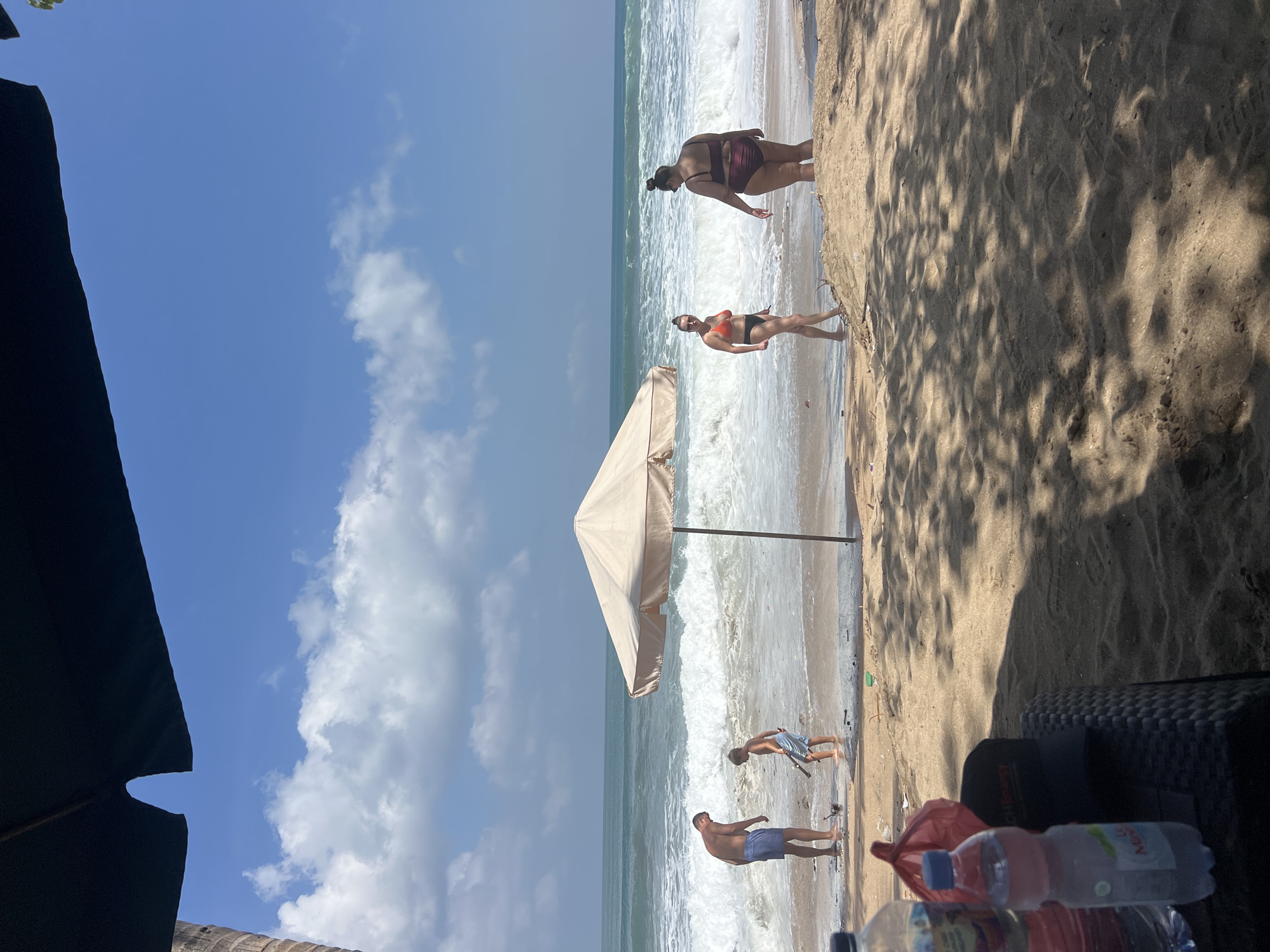 The image captures a sunny day at Kuta Beach, Indonesia. In the foreground, two bottles of water and a red bag rest on a dark woven table, partially shaded by an umbrella. Further back, a family enjoys the ocean. A man in blue shorts and a young boy in light blue shorts are by the water's edge. A large, light-colored umbrella stands between them and the waves. To the right, two women are also near the water; one in an orange bikini and the other in a maroon bikini. The ocean is active with moderate waves and whitecaps, and the sky is blue with scattered clouds. The sand is light brown, and shadows from unseen foliage are cast across it.