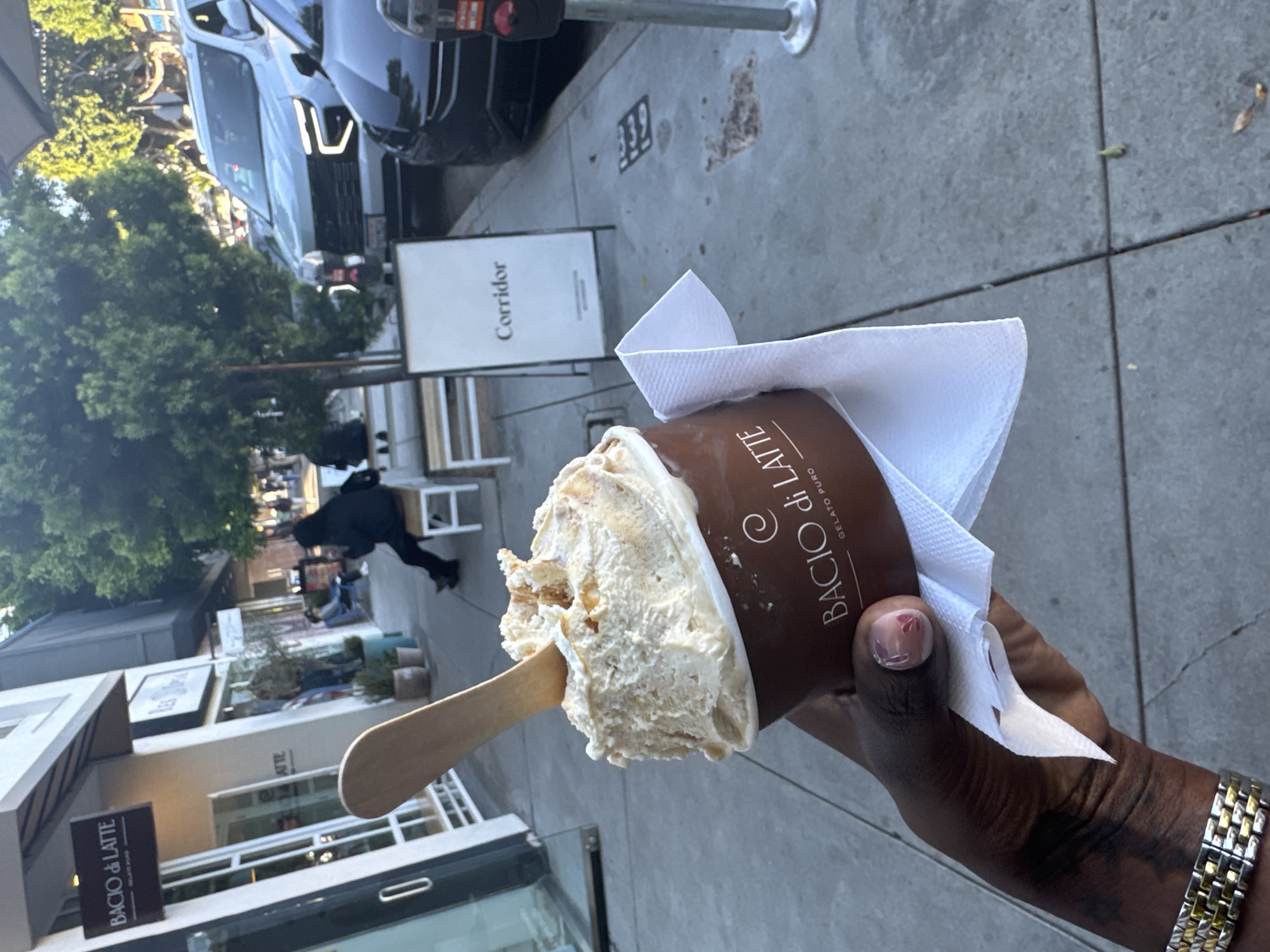 A person stands on a sidewalk, holding a cup of gelato from Bacio di Latte in their right hand. The gelato is scooped onto a wooden stick, and a white napkin is tucked around the cup. The person is wearing a watch with a gold and silver band. The location appears to be a street in Los Angeles, United States of America, based on the Bacio di Latte shop sign in the background and the style of architecture. A "Corridor" sign is also visible. Cars are parked nearby, and people can be seen walking on the sidewalk in the background.