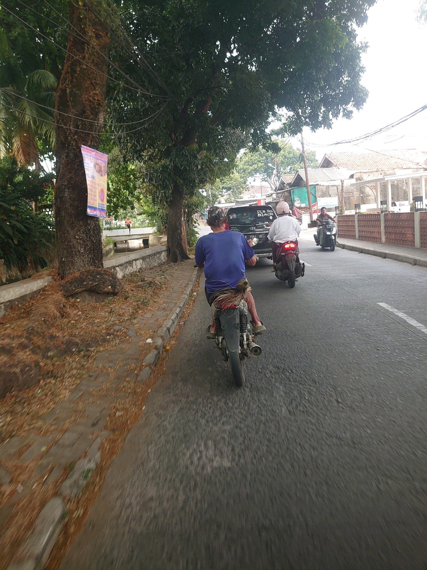 The image depicts a street scene in Depok, Indonesia, likely during daytime given the ambient light. In the foreground, a person wearing a blue shirt and a patterned sarong is riding a motorcycle, facing away from the camera. The motorcycle is silver and black with visible exhaust pipes. Behind this motorcycle, a black car with the text "It's So Black" on its rear window is visible. Another motorcycle is further ahead, to the right of the car, ridden by a person in a white shirt and pink helmet, also facing away from the camera. To the far right, another motorcycle is approaching, with its rider in a dark shirt and helmet.
The street is paved with asphalt, and a sidewalk runs along the left side of the road. The sidewalk is lined with trees, one of which has a poster attached to its trunk. Fallen leaves are scattered on the sidewalk and along the edge of the road. A small canal or water channel is visible between the sidewalk and a low wall, with a bridge crossing it in the background. Buildings with tiled roofs and some with white architectural elements are present in the distance. Power lines are strung across the upper portion of the image. The overall impression is of a typical urban street environment with moderate traffic.