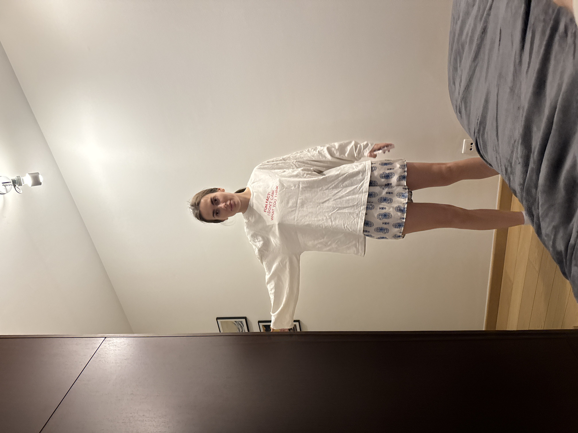 A full-length, sideways photograph captures a woman standing in what appears to be a bedroom. The woman is wearing a white, long-sleeved shirt with red text on the front and patterned shorts. The room features a light-colored wall with mounted light fixtures and framed pictures, a wardrobe with a dark wood finish, and a bed with gray bedding. The location of the image is Unknown, Unknown.