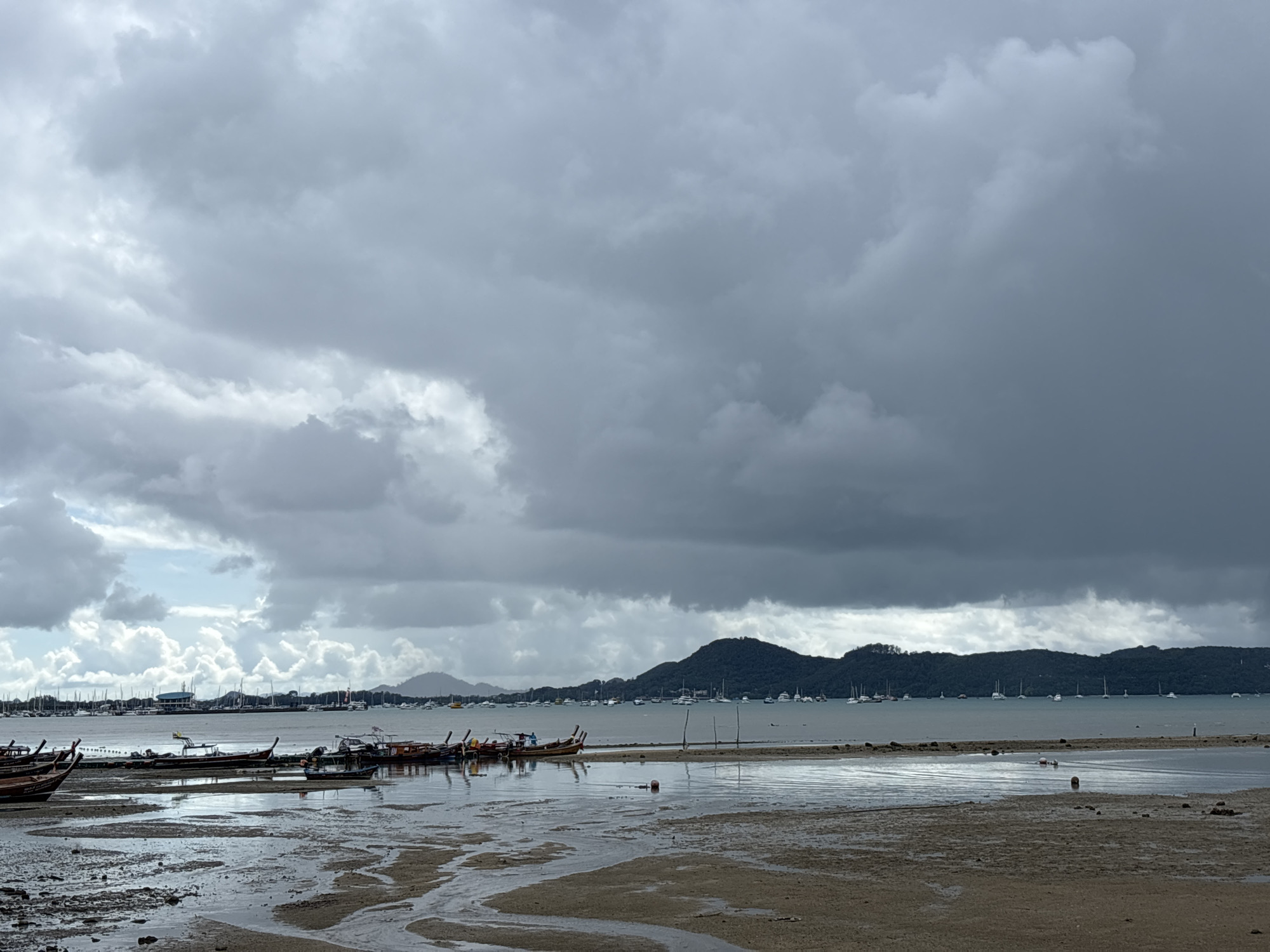 The image captures a cloudy day at the beach in Rawai, Thailand. The foreground is dominated by wet sand and tidal pools, reflecting the overcast sky. A collection of traditional Thai longtail boats are moored on the shallow water and partially on the sand. In the background, the sea is dotted with numerous sailboats anchored in the distance. Beyond the water, a line of lush, green hills forms the horizon. The sky is filled with heavy, dark grey clouds, suggesting an impending storm or recent rainfall, with patches of lighter grey and white clouds breaking through in places. The overall scene conveys a sense of calm before or after a weather event, with the boats resting and the landscape subdued under the dramatic sky.