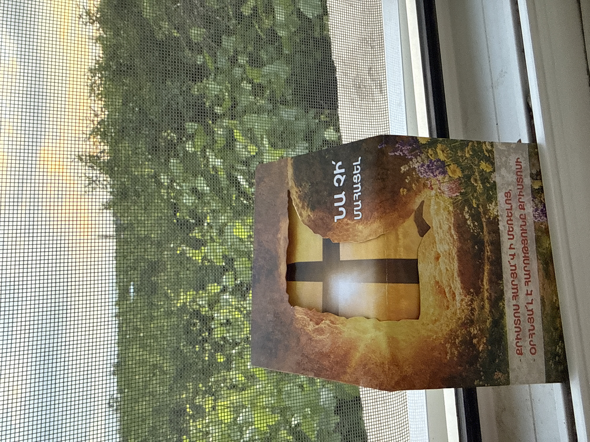 A rectangular, vertically oriented card or small box, featuring a religious theme, is positioned in front of a window. The object has a torn or distressed opening on its front, revealing a glowing cross symbol within, rendered in warm yellow and orange tones, suggesting light emanating from it. The object's design incorporates earthy browns, with hints of purple and green floral elements visible on its right side.
Outside the window, a fine mesh screen is visible, beyond which lush green foliage, likely trees or bushes, can be seen. In the upper left portion of the outdoor view, a bright, light-colored sky suggests a sunny daytime setting.
Armenian text is visible on the object. On the upper left, two lines of text read: "ԱՄ ՁԻՆ" followed by "ՄԱՉԱՑԵԼ". Along the right edge of the object, oriented vertically and read from bottom to top, is the phrase: "ՔՐԻՍՏՈՍՈՒՆ ԶԱՐԳԱՅԻՆ Ի ՄԵՐԵԱԼՈՒՆ. ՕՐԵԱՆՍՆ Է ՀԱՐՈՒԹՅՈՒՆԸ ՔՐԻՍՏՈՍԻ." This phrase is a variation of the traditional Armenian Easter greeting, "Christ is risen from the dead. Blessed is the Resurrection of Christ." No people are visible in the image.