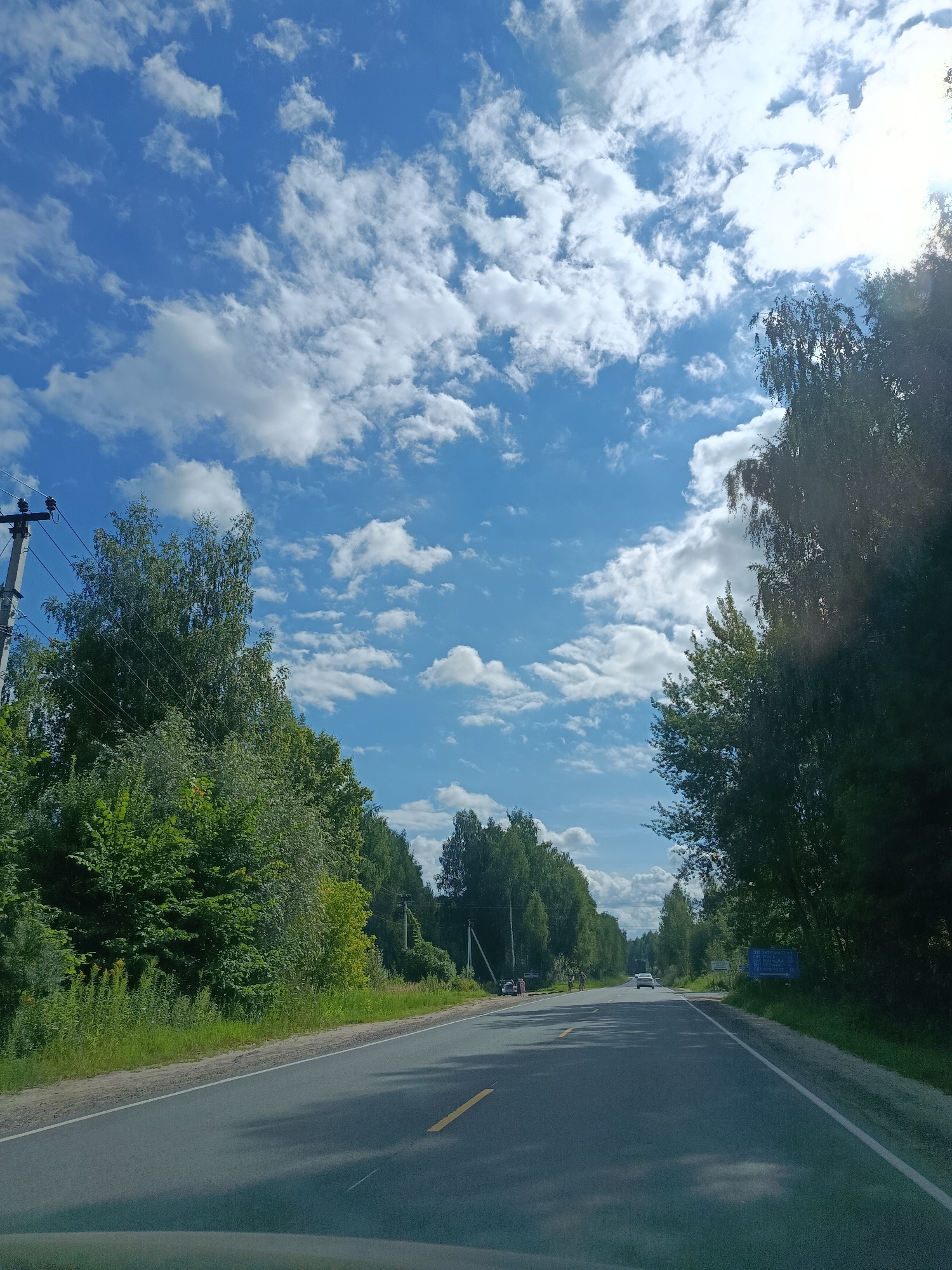 The image depicts a straight, asphalt road with a yellow centerline, bordered by lush green trees and vegetation on both sides. The sky is a vibrant blue, dotted with fluffy white clouds. A utility pole with wires is visible on the left side of the road, and further down the road, several cars and a few people can be seen. A blue sign with white text is posted on the right side of the road, indicating distances to various locations. The scene suggests a sunny day in a rural or semi-rural setting. The image appears to be taken from a moving vehicle, as evidenced by the perspective and slight motion blur. There is no discernible information within the image that places it specifically in the city of 1-Go Maya, Russia.