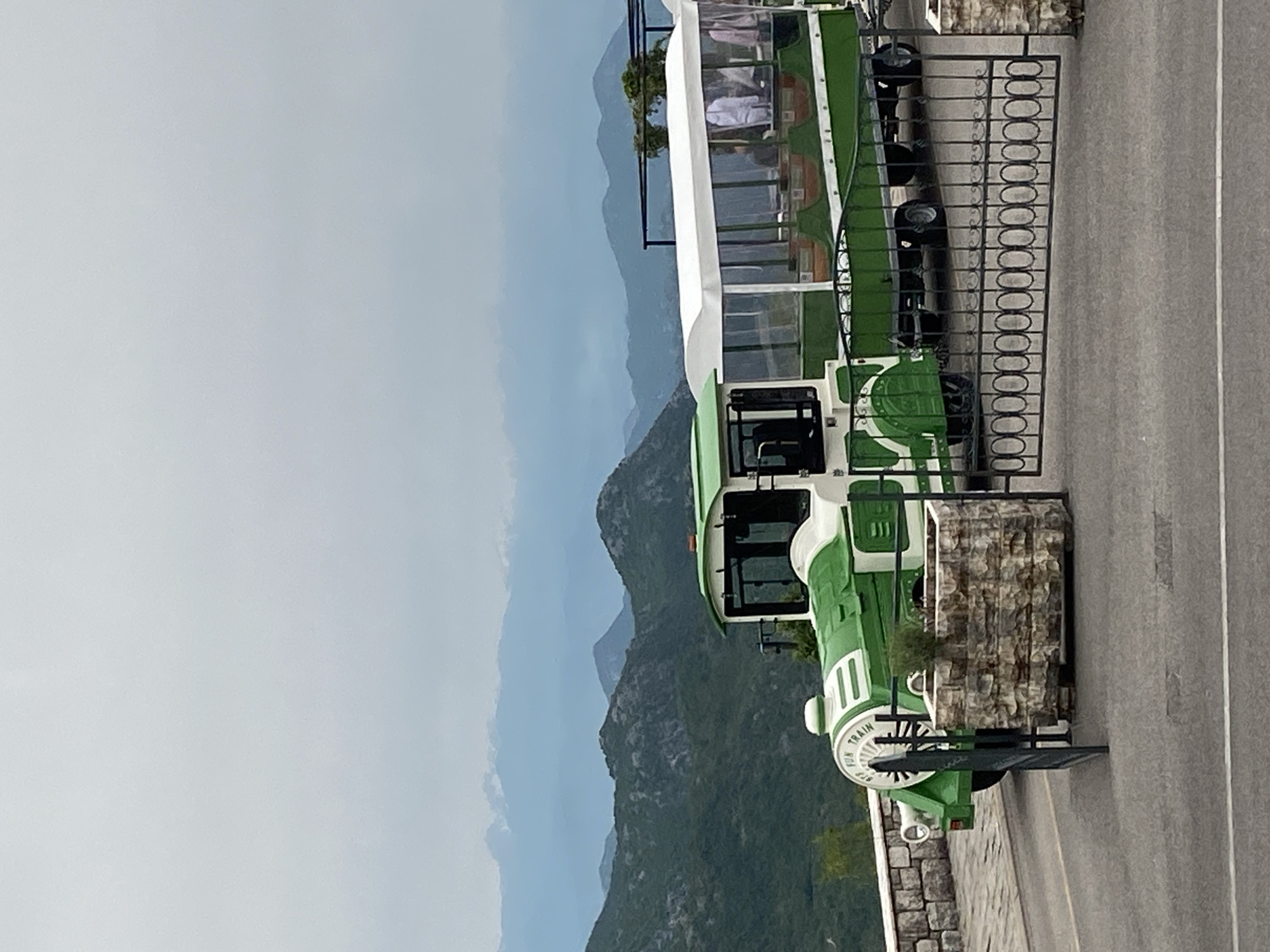 The image presents a green and white tourist train parked next to a metal fence and a paved road. A landscape of mountains and a cloudy sky forms the backdrop. The location is suggested to be Lipa Dobrska, Montenegro, judging by the mountainous landscape.