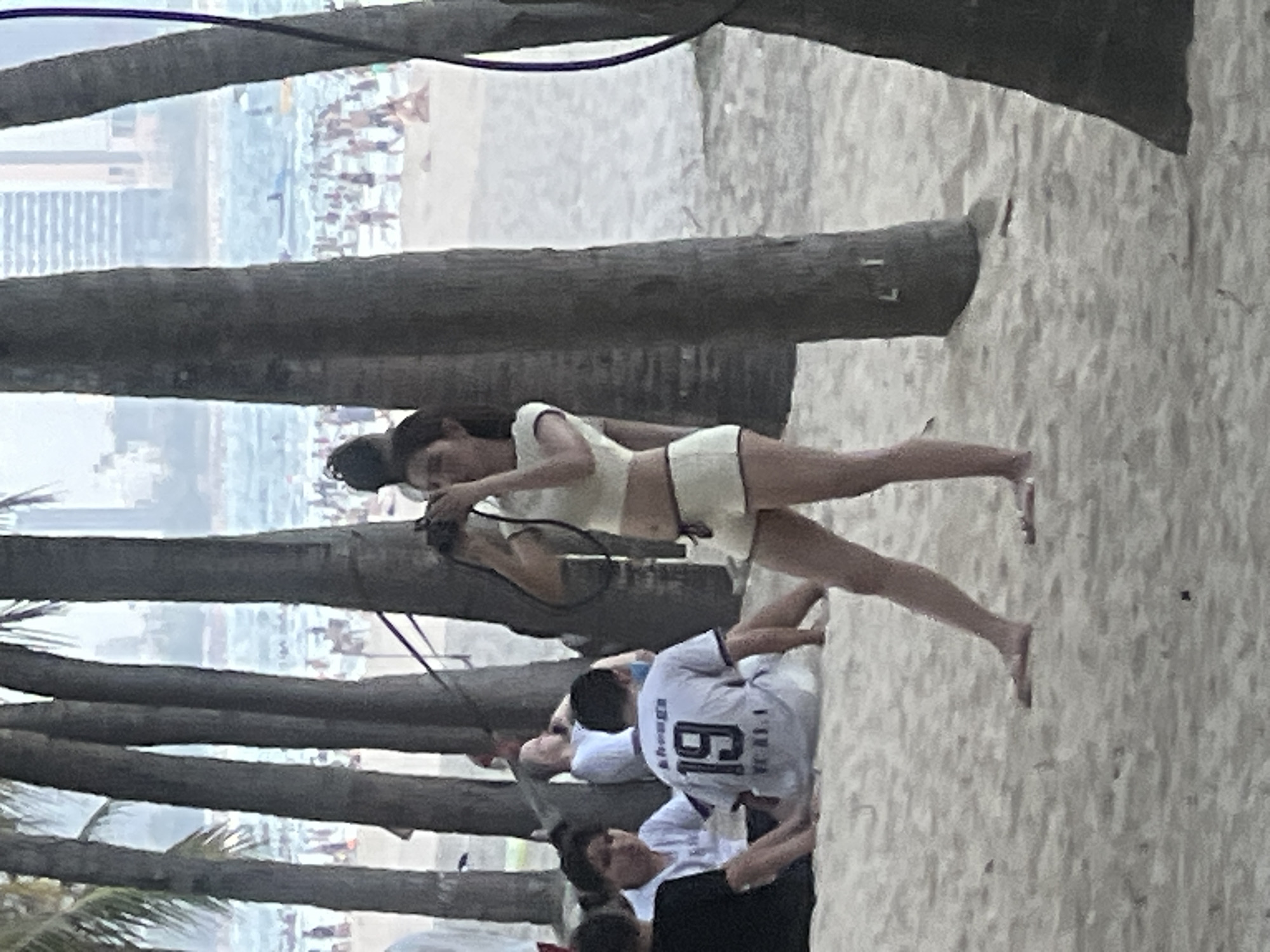 The image depicts a beach scene in Đà Nẵng, Vietnam. The foreground is dominated by the trunks of several palm trees, obscuring parts of the background.
In the middle ground, a woman is standing on the sand, facing towards the left of the frame. She is wearing a light-colored crop top and shorts and appears to be holding a camera. Her body is angled, with her legs extended.
Further in the background, beyond the palm trees, a crowded beach is visible. Numerous people are scattered across the sand and the edge of the water. There are buildings and structures in the far distance, suggesting an urban coastline.
Below the woman, other people are present on the beach. One individual is wearing a white t-shirt with the number "19" prominently displayed on the back. The overall scene suggests a bright day, likely daytime, with moderate activity on the beach. The presence of palm trees and the distant urban landscape are strong indicators of the specified location.