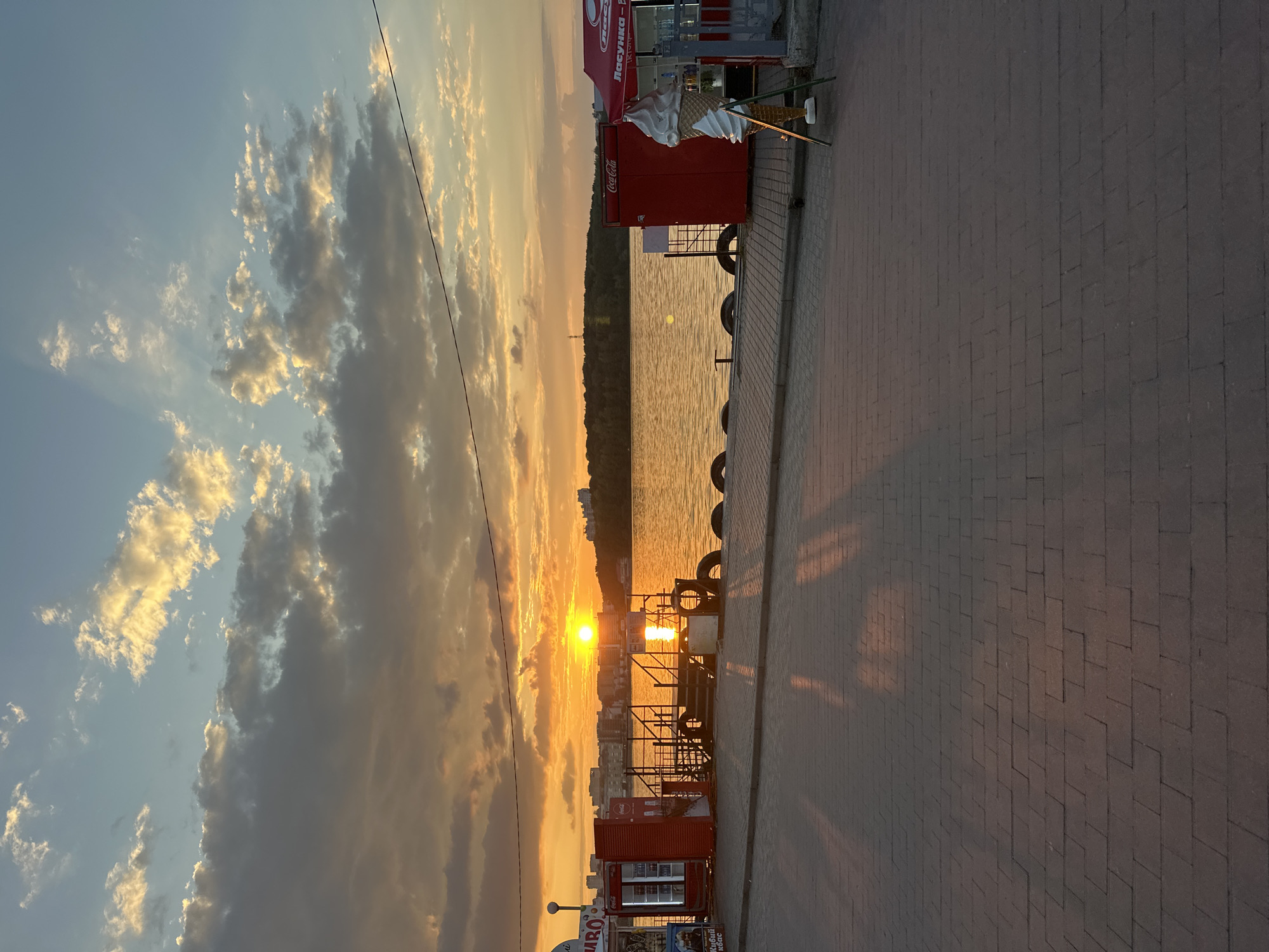 The photograph captures a waterfront scene at sunset in Ternopil, Ukraine. A bright, orange sun is low on the horizon, casting a warm glow across the sky and reflecting on the water. The sky is filled with dramatic clouds, tinged with yellow and orange, with visible sun rays breaking through. In the foreground, a brick-paved promenade runs along the waterfront. To the left, there is a red kiosk with glass doors, likely a food or drink vendor. Further along the promenade, a metal structure with stairs leads down to the water, and several large tires are stacked along the edge of the promenade. To the right, another red kiosk with an umbrella is visible, and a large, artificial ice cream cone sculpture stands on display. The water stretches towards a treeline on the opposite bank, with buildings visible in the distance. The overall impression is a picturesque evening view of a city waterfront.