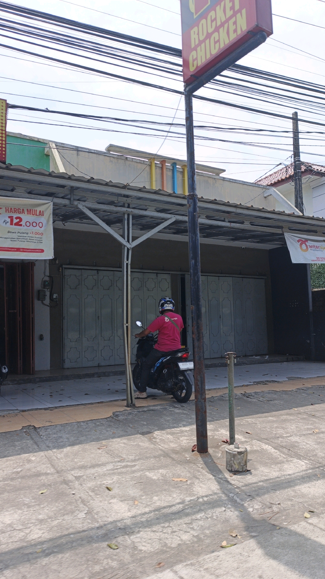 A person wearing a pink shirt and a blue helmet is riding a black motorcycle in front of a building with rolled-down shutters. A sign for "ROCKET CHICKEN" is visible above the building. A banner advertising "HARGA MULAI RP 12.000" is hanging on the left side of the building. The scene appears to be on a street in Desa Wonoharjo, Indonesia, with overhead power lines and a sidewalk in the foreground.