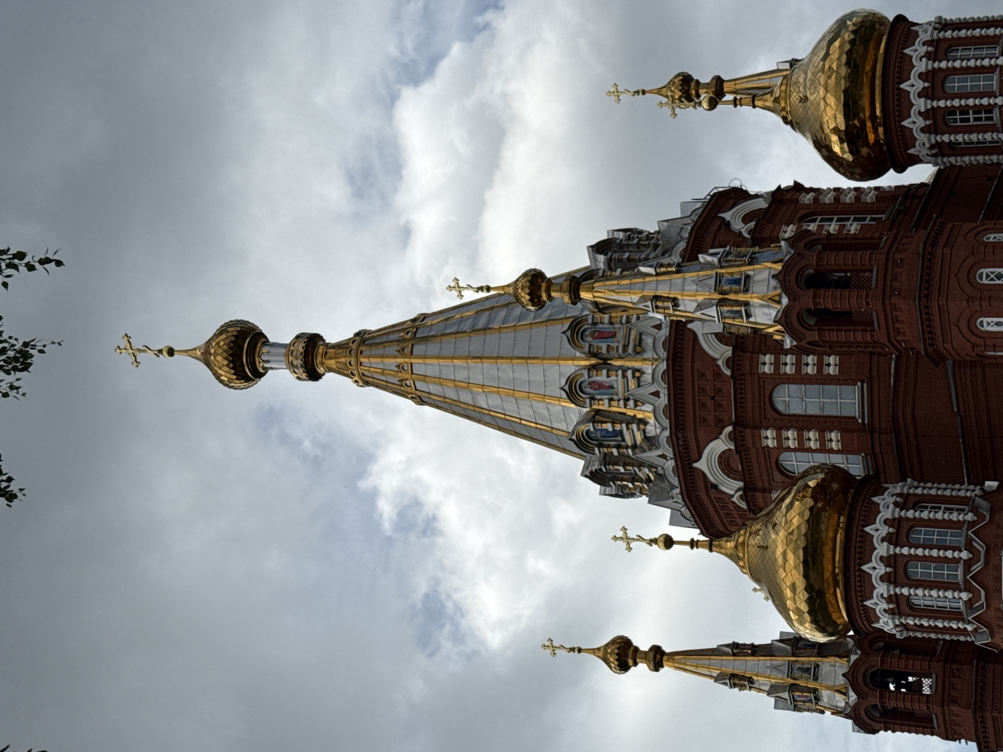 The image shows the domes and spires of the Holy Trinity Cathedral in Izhevsk, Russia, against a cloudy sky. The cathedral is constructed of red brick and features multiple golden domes and ornate architectural details. The uppermost dome has a cross at its apex. There are no people visible in the image. The environment is overcast with some white clouds breaking through the gray.