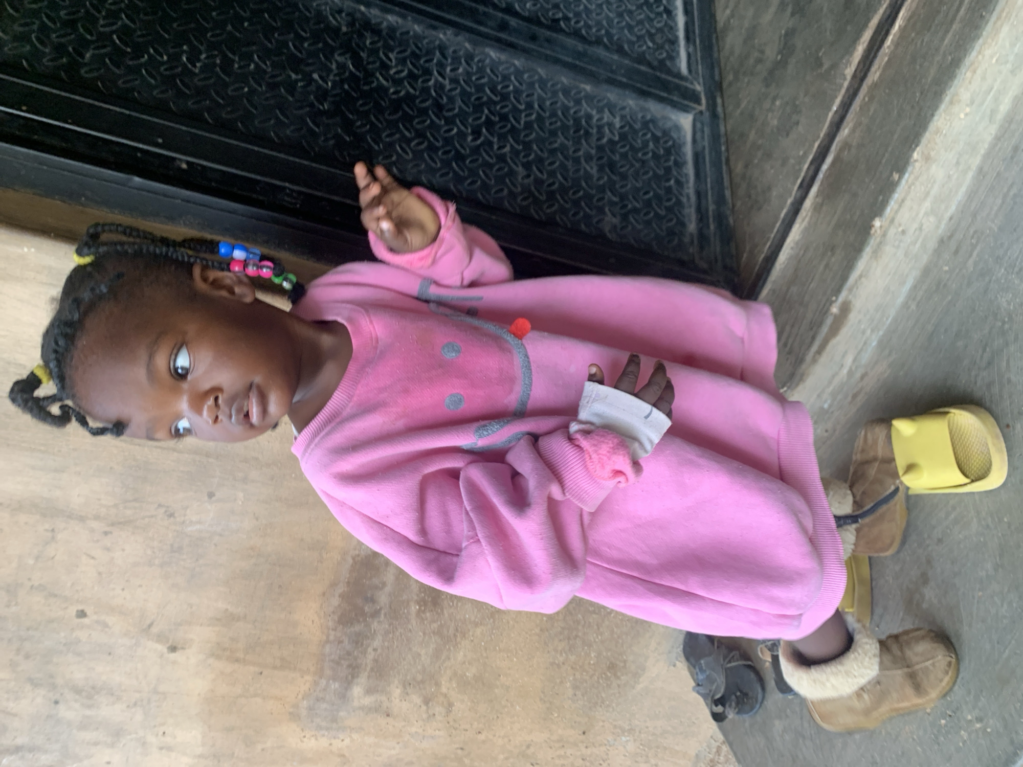 A young Black girl with braided hair adorned with colorful beads stands near a dark metal door. She is wearing a pink sweatshirt with a smiley face graphic and white cuffs on her sleeves. The girl is looking to her right, away from the camera. She is standing on a concrete surface, with several pairs of shoes scattered around her feet, including brown boots and yellow slides. The background consists of a textured, light-colored wall. The scene appears to be outdoors in an urban environment, possibly in the city of Jos, Nigeria, given the architecture and footwear.