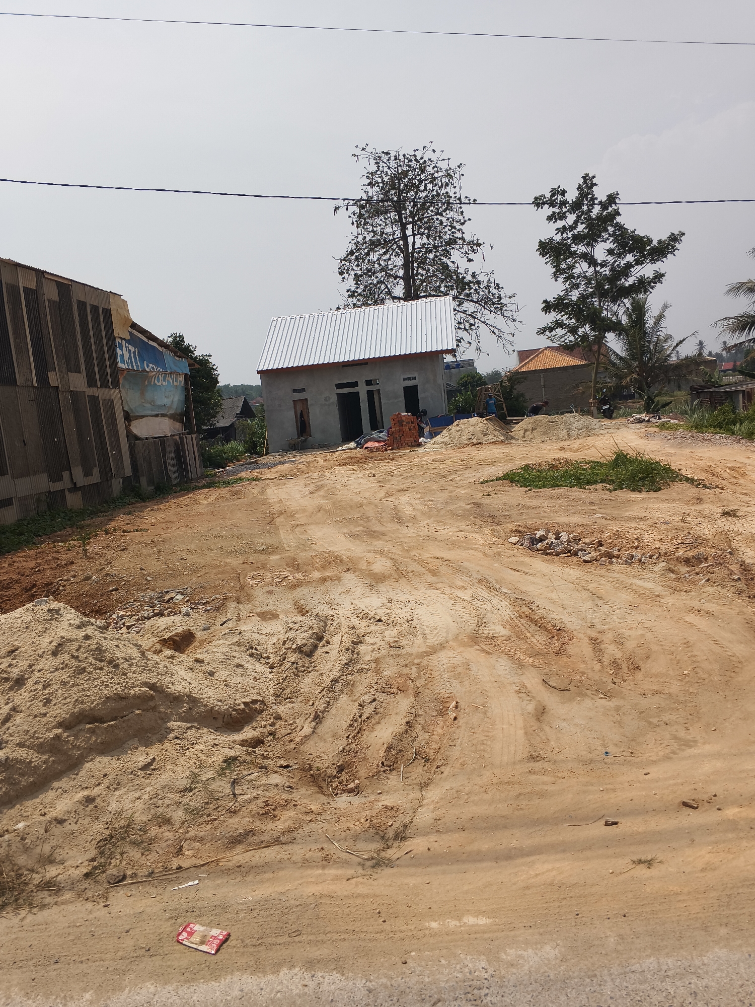 The image depicts an outdoor scene in Banjar Agung, Indonesia, characterized by a construction site or undeveloped land. In the foreground, a dirt path with visible tire tracks dominates the scene, leading towards the middle ground. To the left, a pile of light-colored sand is situated.
In the middle ground, a partially constructed building with a corrugated metal roof is visible. Bricks are stacked near this building, suggesting ongoing construction. To the left of the building, a structure with a corrugated metal wall and a blue and white sign is present. Several trees are scattered throughout the scene, with one notably bare tree standing behind the partially constructed building and a palm tree to the right. Further in the background, other buildings and vegetation are visible.
The sky is overcast and light in color. The overall environment suggests a rural or semi-rural setting undergoing development or preparation for construction. There are no people clearly identifiable in the image.