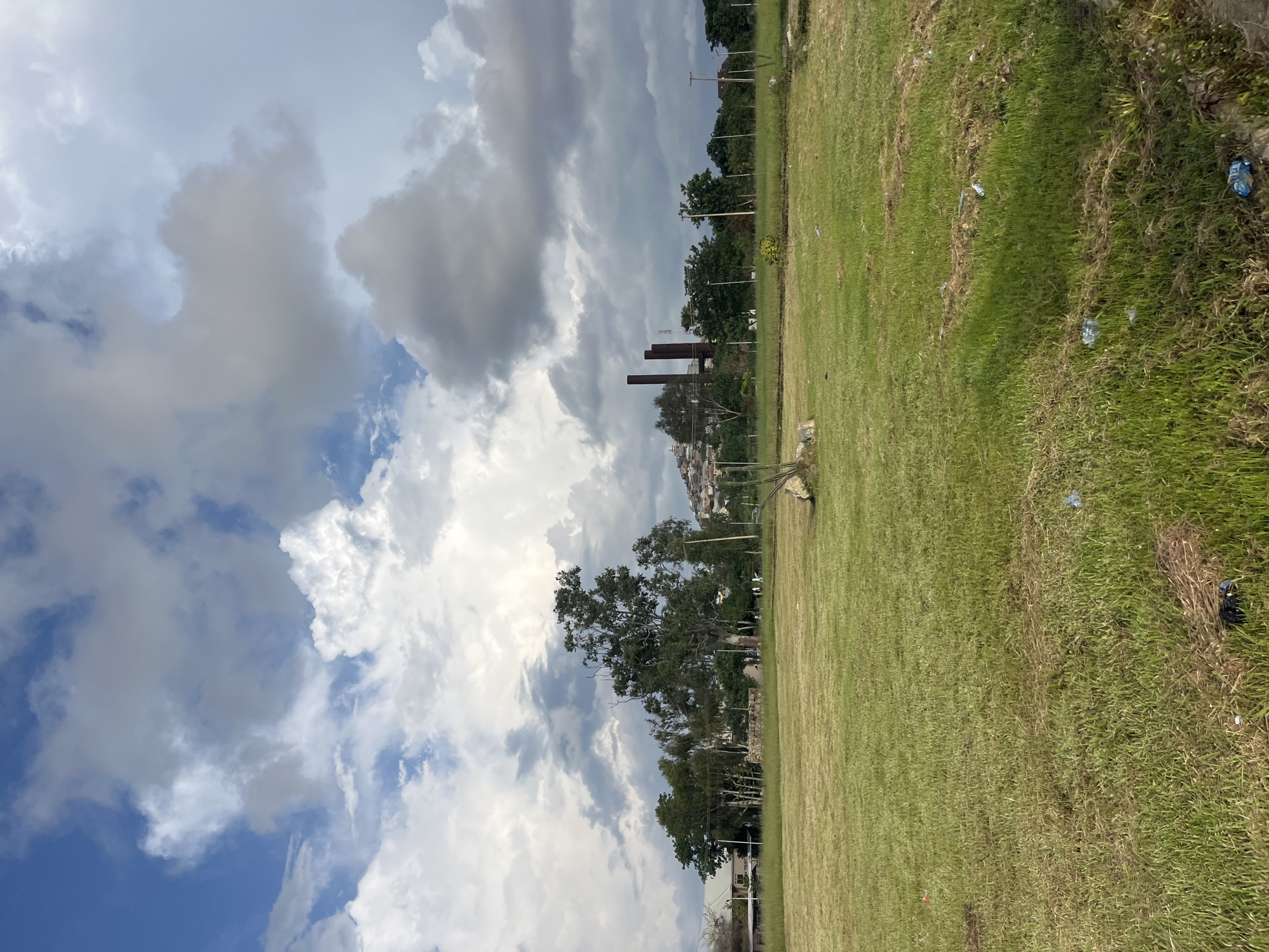 A wide, grassy field occupies the foreground of this photograph, appearing slightly overgrown in places with patches of dry grass and scattered debris, including plastic bags and other litter. In the middle ground, a cluster of trees stands on a gentle rise. To the left of the trees, a light-colored building with a sloped roof is visible. Further back and to the right, a line of several tall, rusty smokestacks dominates the skyline, suggesting an industrial site. Behind the smokestacks, a densely packed hillside settlement with buildings is visible, characteristic of areas such as the Angulap Quarter or the Dogo Na Hauwa area in Jos, Nigeria, which are known for their informal housing on elevated terrain and proximity to former industrial areas. The sky above is filled with a dramatic display of clouds, with patches of blue sky peeking through. The overall scene depicts a partially developed or transitioning landscape within the urban environment of Jos.
