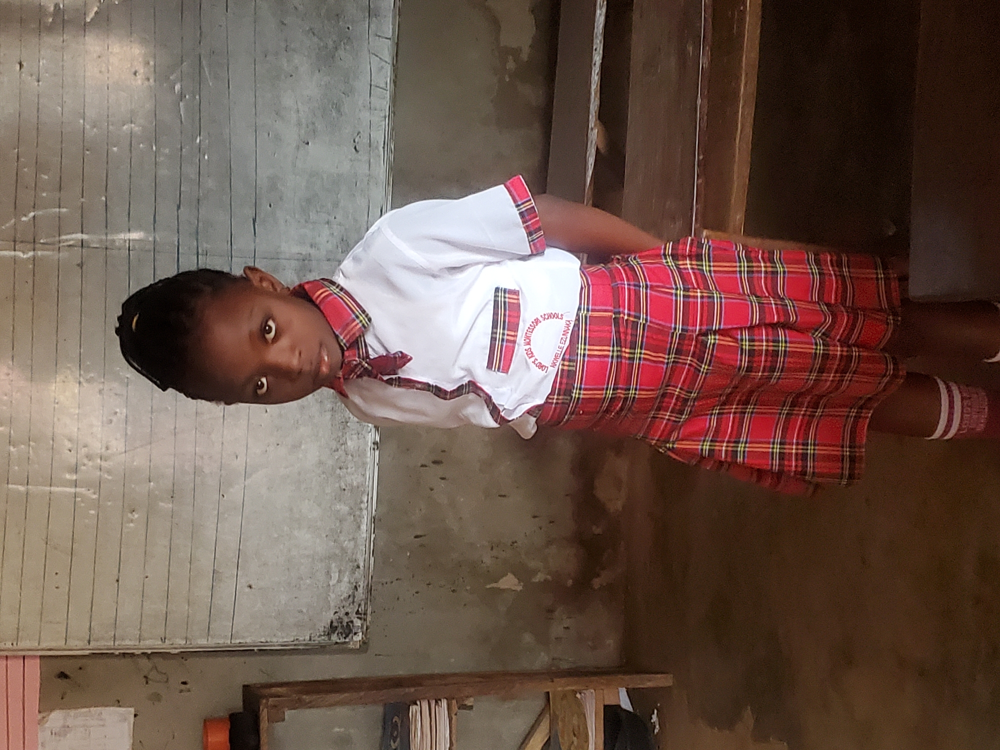A young girl stands in what appears to be a classroom in Ukwu-Abwa, Nigeria. She is wearing a red plaid school uniform with a white shirt bearing the name of her school. The room features a writing board, and assorted items are stored on a wooden shelf. The walls exhibit signs of wear and discoloration.