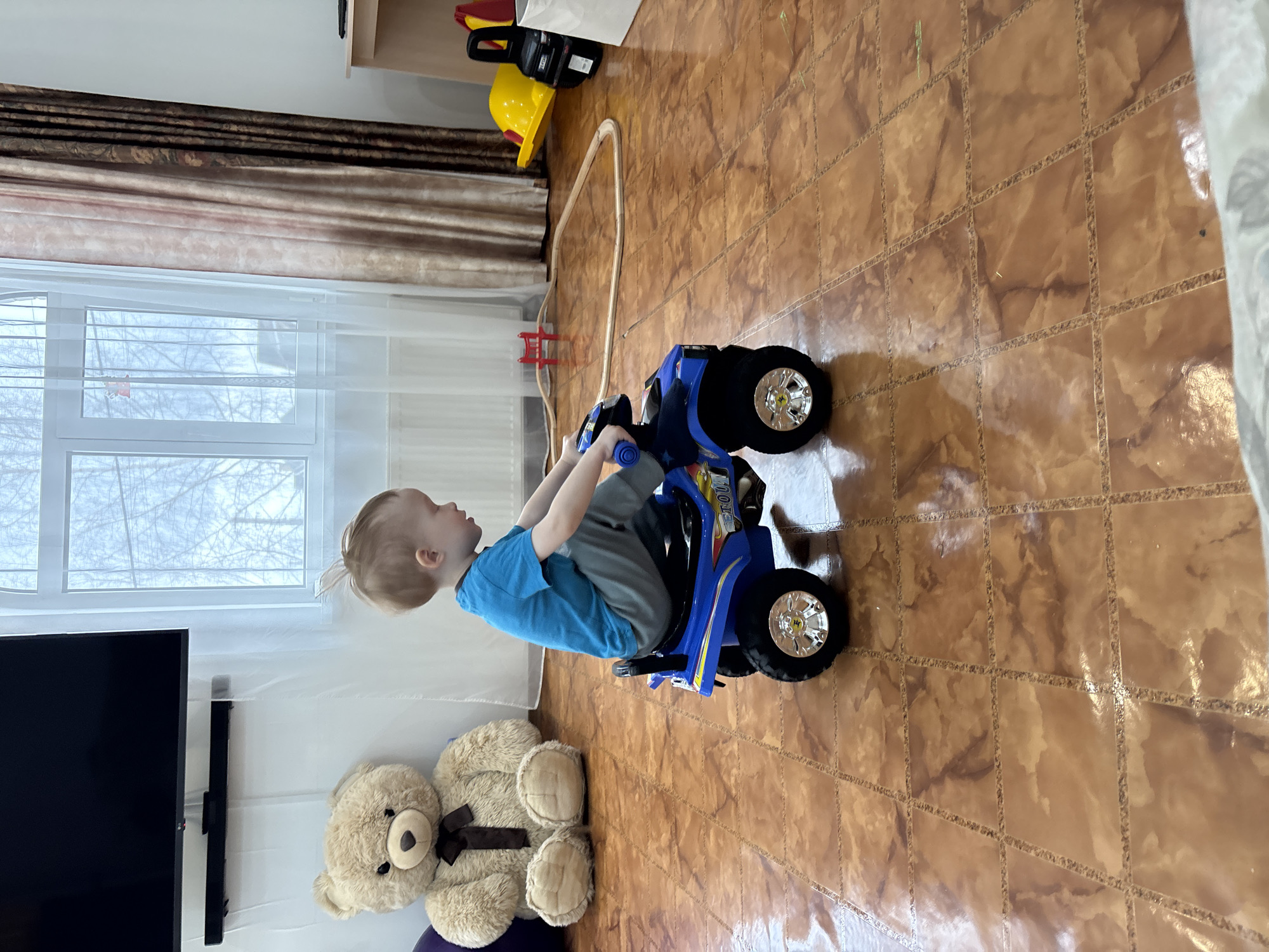 The image depicts a young child sitting on a blue toy ATV in what appears to be an indoor setting. The child is fair-skinned with blond hair, wearing a blue t-shirt and grey pants. The child is facing towards the right side of the image.
The setting is a room with brown tiled flooring. In the background, there is a window covered with white curtains and brown draperies, a black television mounted on a wall, and a large teddy bear. Near the window, there's also a toy train set and a yellow construction hat on a shelf. The location may be inferred as the city of Ферма, Russia, based on the user's request.