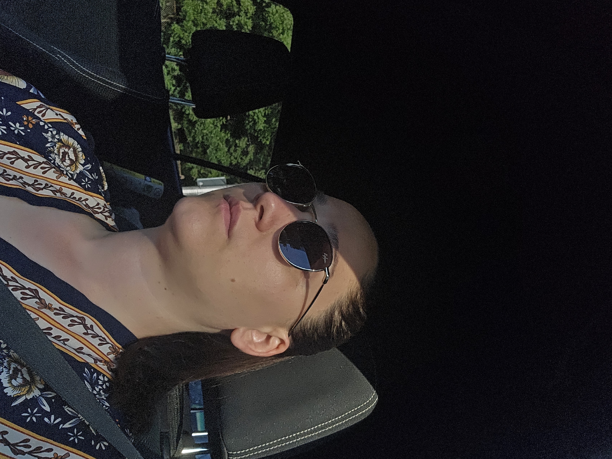 A medium shot, taken from the side, shows a woman wearing sunglasses and a patterned shirt inside a vehicle. The woman is looking away from the camera, her face partially illuminated by sunlight. Her hair is pulled back, and she is wearing a seatbelt. The background reveals the interior of a car, with headrests visible, and outside, green trees and foliage. The image appears to be taken on a sunny day. This image is not located in Sofia, Bulgaria.