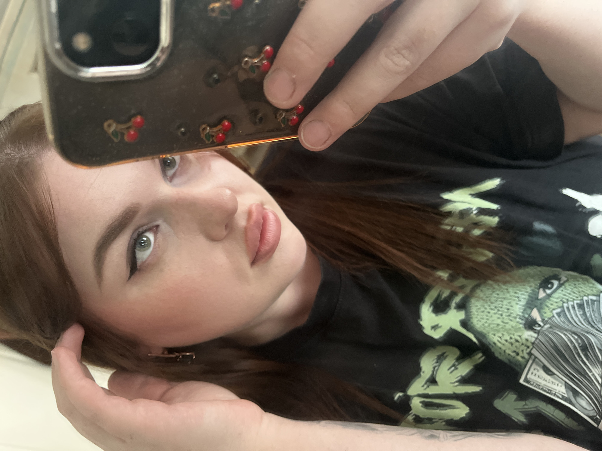 This close-up selfie, taken in Pavlovka, Russia, features a young woman holding a smartphone, likely reflecting her image. The woman has fair skin, reddish-brown hair, and light blue or grey eyes with visible winged eyeliner. Her lips are slightly parted, revealing a light pink lip color, and she has a hint of blush on her cheeks. She is wearing a black t-shirt with a prominent graphic design.
The design on her t-shirt includes a creature, possibly a bird or a monster, rendered in shades of light green. This creature has distinct eyes and appears to have feathers or a body composed of stacks of dollar bills, which are clearly visible on the right side of the graphic. Other abstract green shapes and stylized elements, including what looks like a "V" shape, are also part of the design.
The woman is holding a dark-colored smartphone with a transparent or dark case adorned with small, red cherry embellishments. Her fingers are visible on the phone case, and her other hand is near her hair on the left side of the frame. The background is mostly out of focus or obscured by her hair and body, suggesting a simple, light-colored indoor setting. The overall mood of the image is calm and posed.