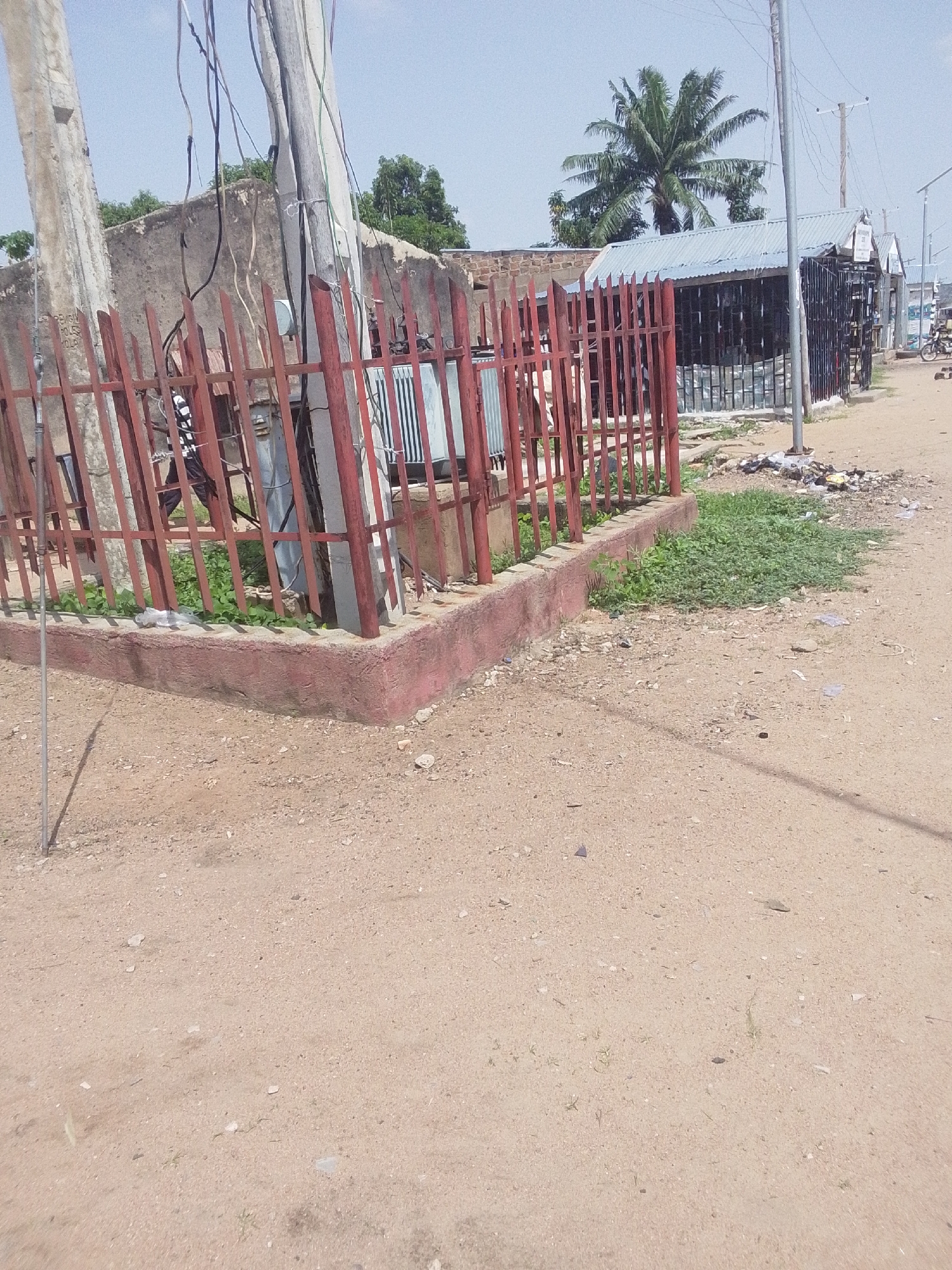 This image depicts a street scene in Jalingo, Nigeria. In the foreground, a dirt road is visible, with scattered pebbles and dry vegetation. To the left, a red metal fence encloses a small area with a concrete base. Behind the fence, a large electrical transformer is situated next to a concrete utility pole with numerous dangling wires. Beyond the fence and transformer, a rough, textured building wall can be seen. Further into the background, several small, open-fronted structures with corrugated metal roofs line the street. A tall palm tree stands prominently behind these structures. The sky is clear and blue. The overall impression is of a functional, somewhat cluttered urban environment.