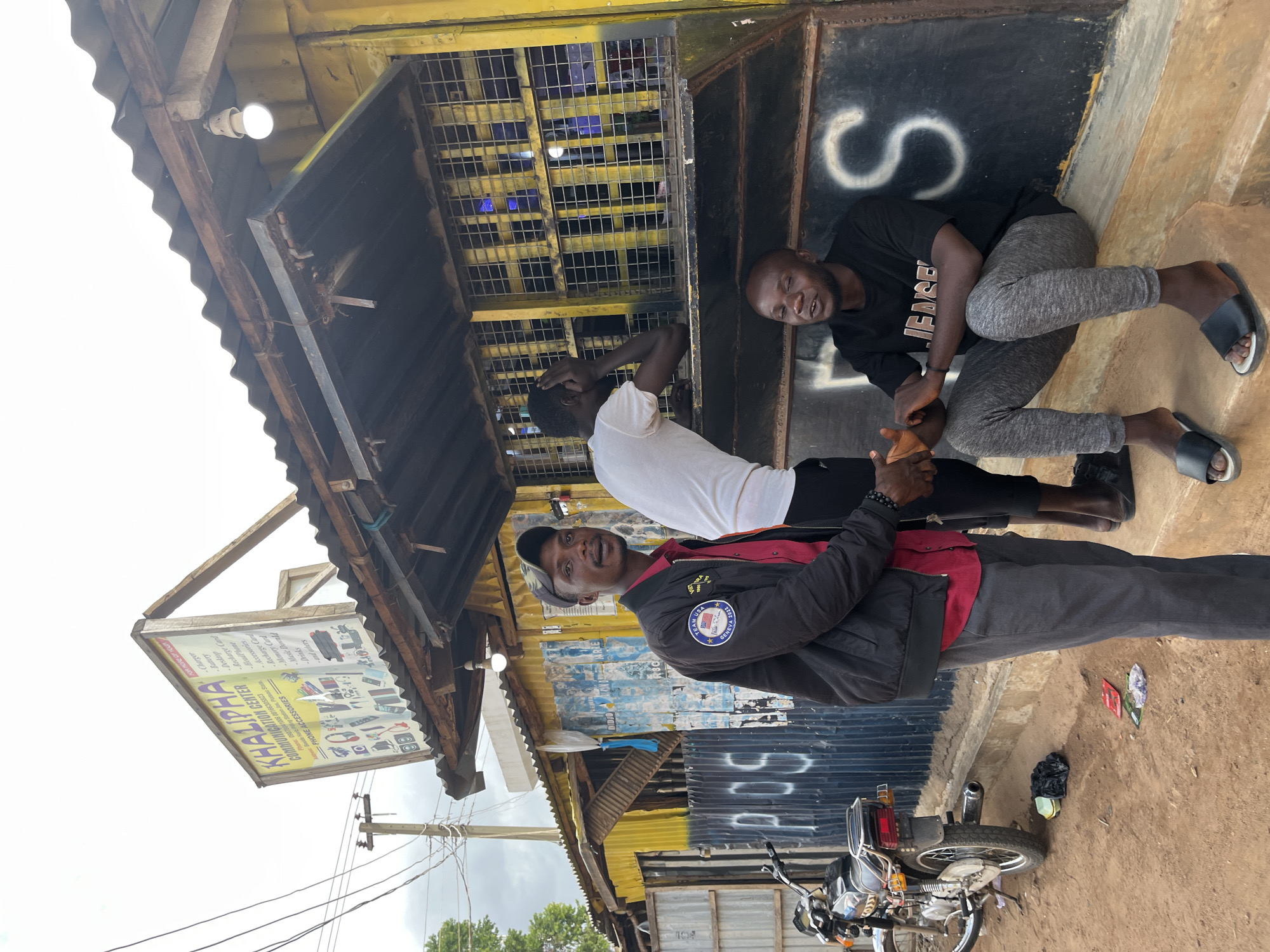 In Rafin Par, Nigeria, three individuals are pictured near a corrugated metal building with a yellow trim, possibly a shop or workshop. The building's facade features black metal doors and a large, white "S" painted on one section. A sign for "KHALIPHA COMMUNICATION CENTER" is visible, advertising phone accessories and providing contact numbers.
Two men are seated near the entrance. One, wearing a black t-shirt and grey sweatpants, is leaning back and smiling, looking towards the camera. The other man, in a white t-shirt, is partially obscured. A third man, wearing a black jacket with an embroidered "TEAM USA GENEVA 2022" patch and a dark baseball cap, stands in the foreground, looking at the camera. He appears to be engaged in a conversation or interaction with someone not fully in frame.
A motorcycle is parked on the dirt ground in front of the building. Various items, including a black plastic bag and some colorful packets, are scattered on the ground near the motorcycle. The lighting suggests it is daytime, and the scene appears casual and outdoors. The overall impression is a snapshot of daily life in a commercial area, with people interacting and showcasing goods or services.