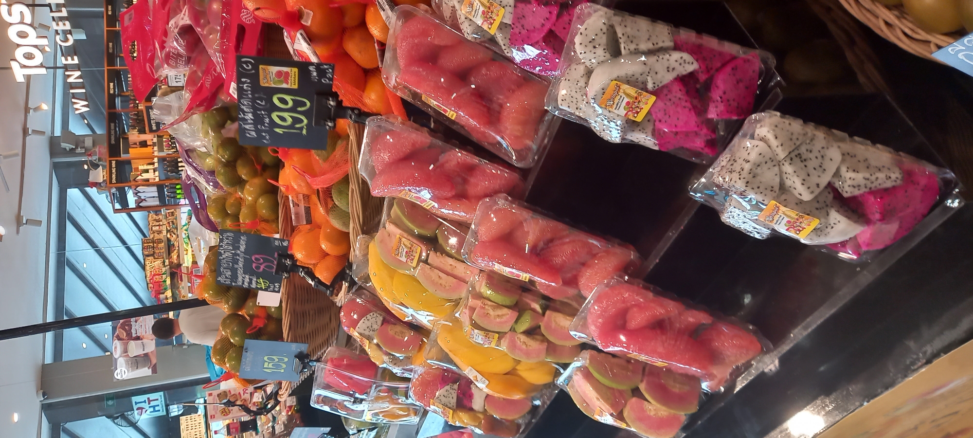 The image captures a display of packaged and unpackaged fruits in what appears to be a supermarket setting in Thailand, specifically in the city of Moo 1. On the left, one can see the signage for "Tops WINE CELL," indicating a wine section nearby, and shoppers are visible in the background. The display prominently features a variety of fruits, including unpackaged oranges and mandarin oranges in baskets, and individually packaged cut fruits. The cut fruits, sealed in clear plastic, include watermelon slices, guava slices, and dragon fruit, distinguished by its pink and white flesh with black seeds. Price tags in Thai currency are visible, with one indicating a price of 89 and another at 199. The scene suggests a supermarket aisle featuring fresh produce in a commercial environment in Thailand.