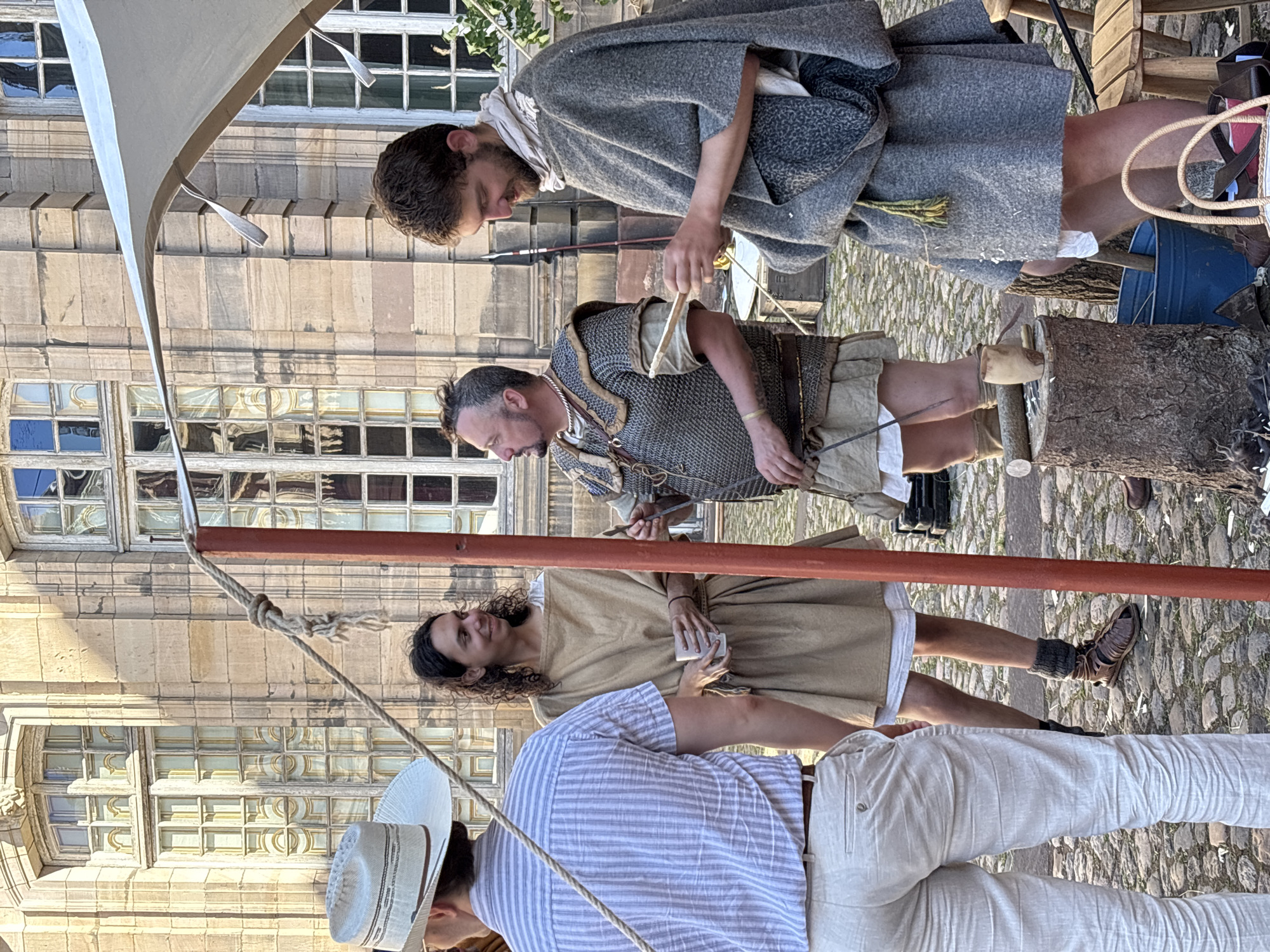 This image depicts an outdoor scene in front of a building with classical architecture, likely in Strasbourg, France, given the style of the building and the cobblestone ground. Four individuals are present, all appearing to be reenactors or participants in a historical event.
On the left, a person wearing a light-colored striped shirt and light-colored trousers stands with their back to the camera, facing towards the right. They are wearing a light-colored wide-brimmed hat.
In the center, a person with curly dark hair is wearing a tan tunic and holding a white cup. They are looking to their left, with a slight smile. Beside them to the right, another individual is dressed in what appears to be Roman military attire, including a chainmail shirt over a tunic and a leather belt. They are holding a tablet-like object and looking to their left.
On the far right, a person with short brown hair and a beard is wearing a grey cloak or tunic over other garments. They are looking down at a piece of paper or tablet they are holding in their hands.
The environment is an outdoor setting with a cobblestone ground. A red-painted wooden pole, part of a temporary structure, is prominently visible in the foreground, running vertically through the frame. There is also a tent or canopy structure visible at the top right of the image. In the background, the building features large, multi-paned windows and stone detailing. Several wooden objects, including a stump with carved items and a blue bucket, are present on the ground.