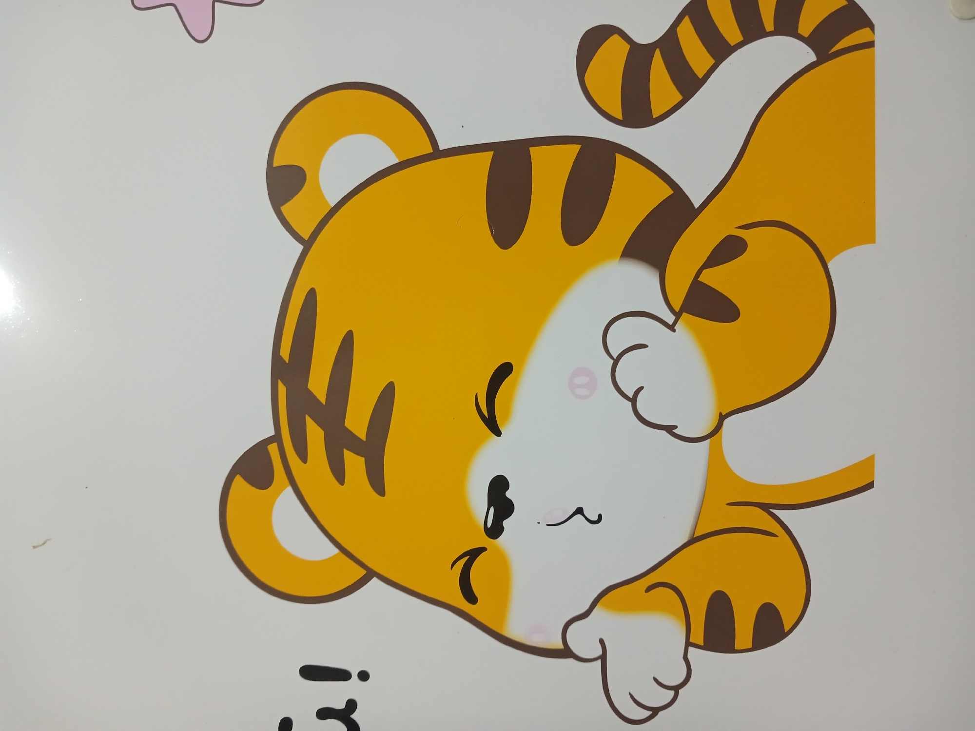 A cartoon illustration of a tiger blowing a kiss is depicted on a white surface, possibly a wall or a piece of furniture. The tiger is predominantly yellow with brown stripes. It has large, round ears, closed eyes with curved lines, and a small, puckered mouth. Its paws are held up to its face as if to blow a kiss. In the upper left corner, there is black text that appears to say "G!" followed by an exclamation mark. To the right of the tiger, a stylized pink star shape is partially visible. The background is a plain white. The image is a close-up, with the tiger filling most of the frame. There is no identifiable context within the image that would place it in Randuagung, Indonesia.