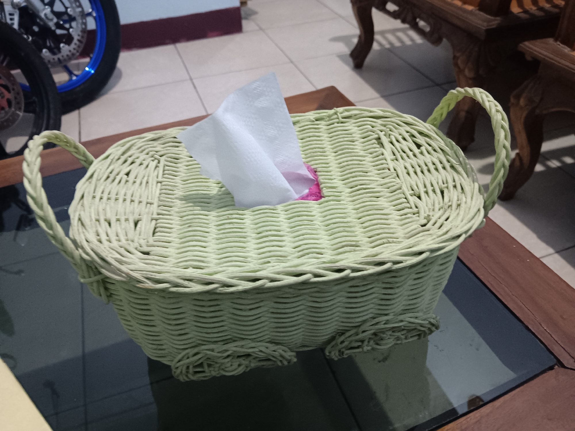 A light green woven basket, filled with white tissues, sits on a dark, reflective surface, likely a glass-topped table. The basket has two braided handles and decorative braided accents at its base, suggesting it is a tissue box cover. Behind the table, a portion of a motorcycle is visible, with its blue rimmed wheel and silver brake disc indicating it is a modern sportbike. To the right of the basket, a wooden ornate chair with carved details is partially in view. The floor is tiled in a neutral color, and in the background, a wall with a dark red and white horizontal stripe is present. The scene is indoors, possibly a residential or common area in Batursari, Indonesia.