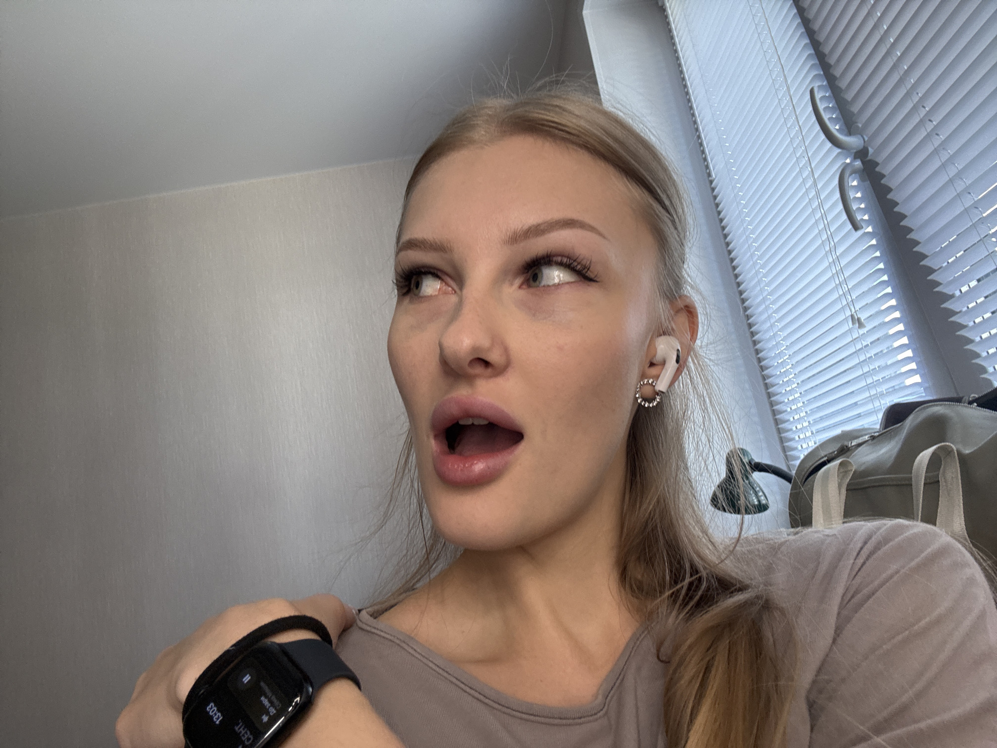 The image shows a young woman with blonde hair, looking to her left with her mouth open in surprise. She is wearing a grey t-shirt, a smartwatch on her left wrist, and white earbuds. The background appears to be an indoor setting with a window covered by white blinds. The overall scene is a close-up of the woman's face and upper body, with a blurred background that includes a wall and a window. There is no visible indication of the city of Perm, Russia, or any specific location.