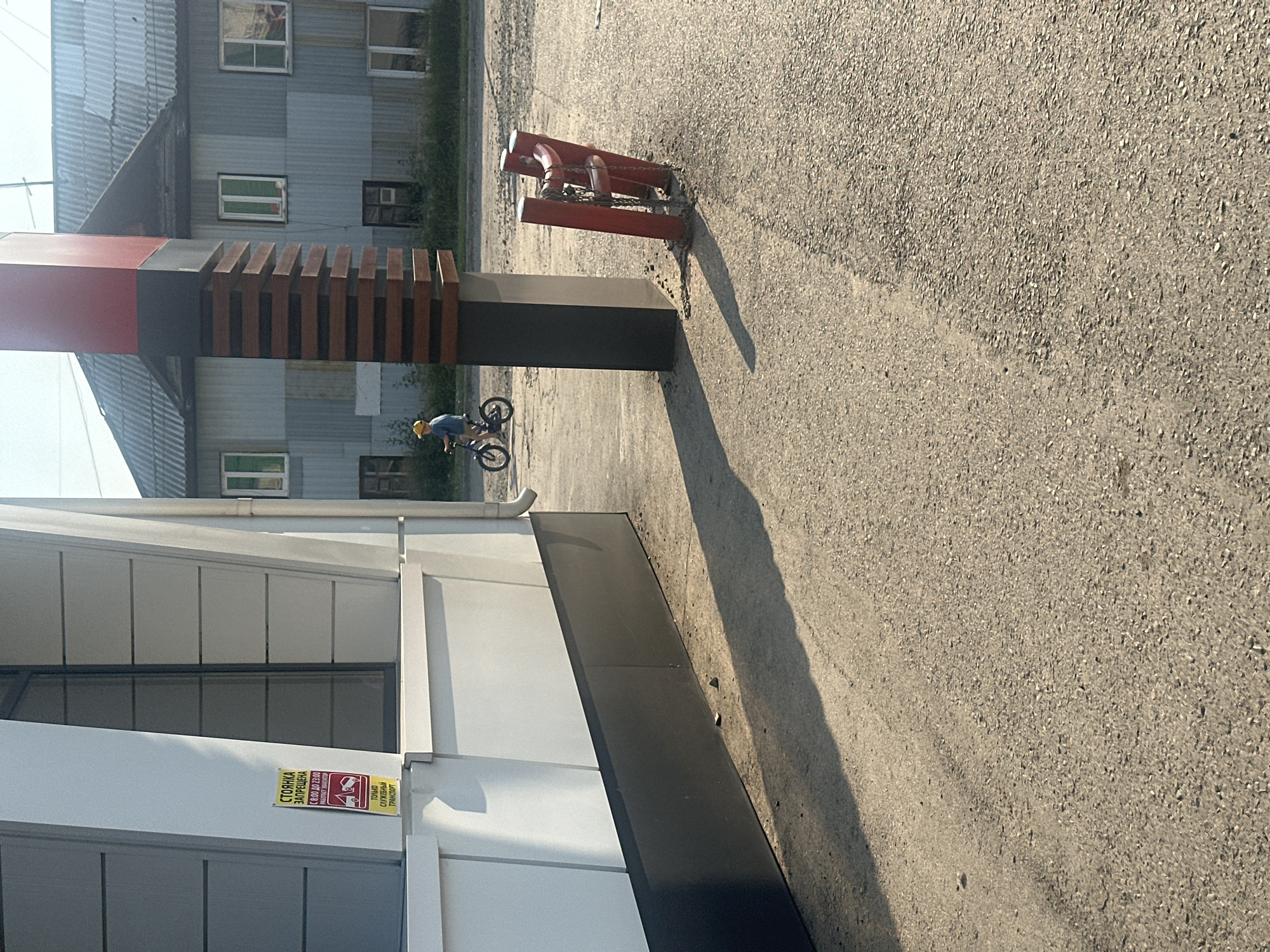 In Novoaltaysk, Russia, a young child wearing a yellow cap and a blue shirt rides a bicycle on an asphalt surface. The child is casting a shadow to the left. Behind the child, a modern building with a white facade and gray horizontal siding stands. A yellow sign with Russian text, indicating parking restrictions and times, is affixed to the building. Further in the background, a dilapidated two-story building with a corrugated metal exterior and worn roof is visible. To the right of the modern building, a black and red vertical structure is present, with wooden slats on its upper section. In front of this structure, red bollards are installed, connected by a metal chain. The overall scene is an outdoor urban environment, likely a street or a public area, illuminated by natural sunlight, creating distinct shadows.
