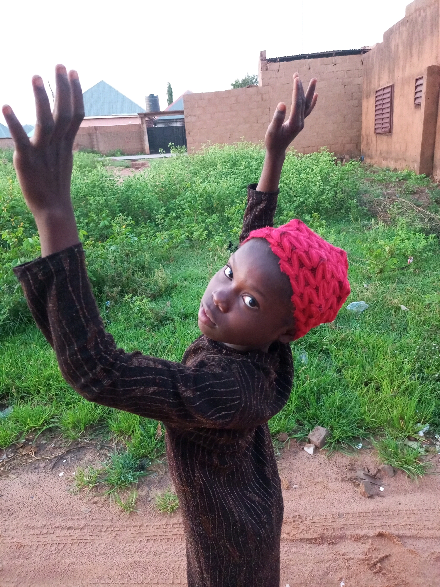A young Black child wearing a brown patterned long-sleeved shirt and a red knitted hat is standing outdoors in what appears to be Tempure, Nigeria. The child is looking towards the camera with arms raised, palms facing outward, and fingers spread. They are positioned in front of a dirt road with tire tracks and some sparse green vegetation. In the background, there are buildings constructed of brown bricks, some with windows, and a water tank. The sky is overcast and bright. The overall setting suggests an informal urban or semi-urban environment.