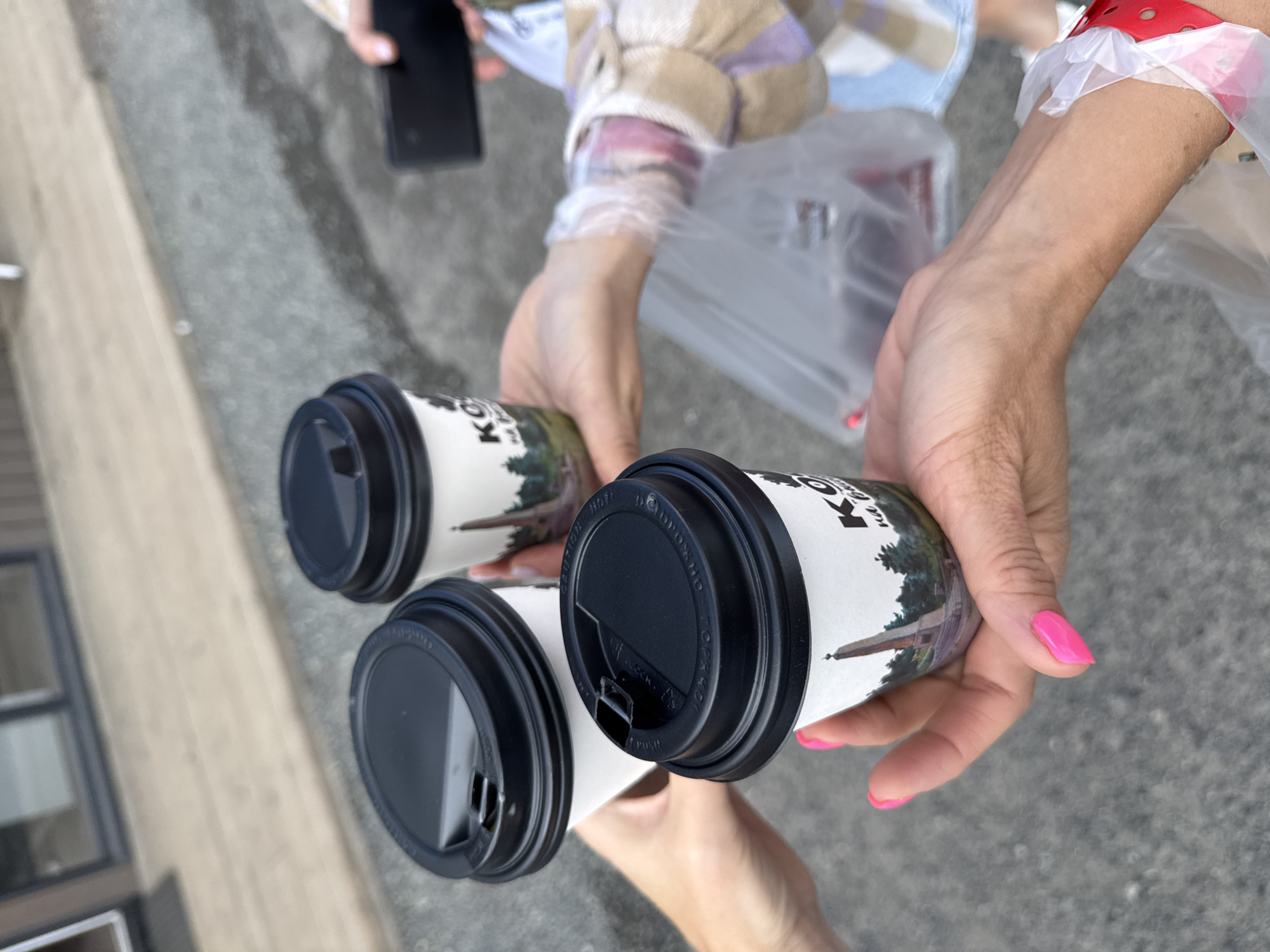Three to-go coffee cups with black lids are held by three individuals, forming a toast. The cups have white paper sleeves featuring a picture of a monument and trees, along with the text "KO". One individual's hand, with bright pink painted nails, is holding a cup in the foreground. To the right, another person's hand is visible holding a smartphone. In the background, a plastic bag with items inside and a person wearing a plaid shirt are partially visible. The scene appears to be outdoors on asphalt, with a building and its entryway visible in the far background. The specific location in the city of T/B Seminskii Pereval, Russia is not identifiable from the visual information present in the image.