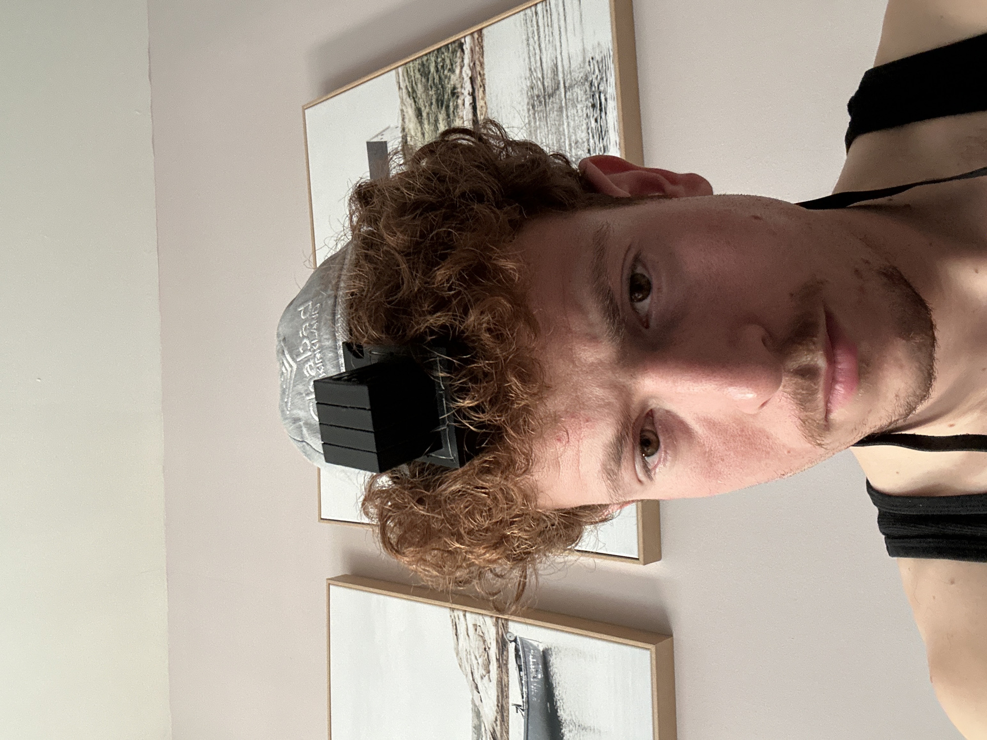 The image presents a close-up, frontal portrait of a young adult male, located in New York City, United States of America. The subject has reddish-brown curly hair, fair skin, brown eyes, a mustache, and light facial hair. He is wearing a black tank top. A black leather cubical object, identified as a *shel rosh* tefillin box, is positioned on his head, centered above his forehead. This object rests on a light-colored, possibly grey, knitted skullcap, which displays text including "Chabad" and "KIRKLAND". A black strap, connected to the tefillin, is visible extending downwards near his right shoulder. The subject gazes directly at the camera with a neutral expression. The background consists of a two-toned interior wall: the upper section is off-white, and the lower section is light pink. Two framed landscape art prints are visible on the lower wall, depicting bodies of water with shorelines and distant terrain.