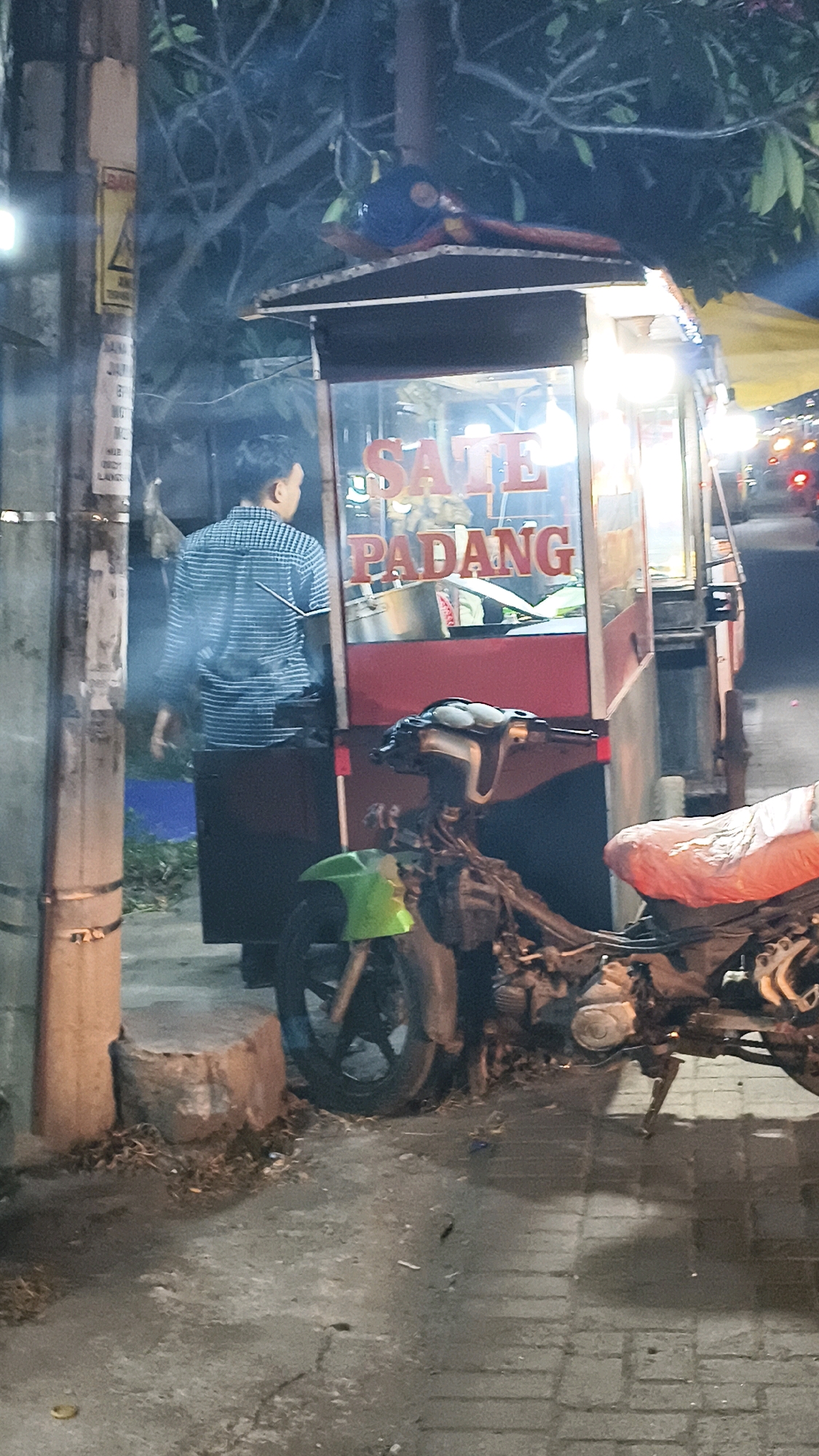 The image captures a nighttime scene in Karangsatria, Indonesia, featuring a food cart that appears to sell "Sate Padang." A person wearing a blue checkered shirt and a dark apron is standing behind the counter of the food cart, facing away from the camera. The cart itself is illuminated, with the words "SATE PADANG" prominently displayed in red lettering on its glass front. In the foreground, a partially dismantled motorcycle is parked. Its front fender is bright green, and the rear seat is covered with a red plastic sheet. The overall environment suggests a street-side setting, possibly a marketplace or a roadside stall, with a pole and some dry leaves visible on the ground. The lighting is dim, indicative of nighttime.