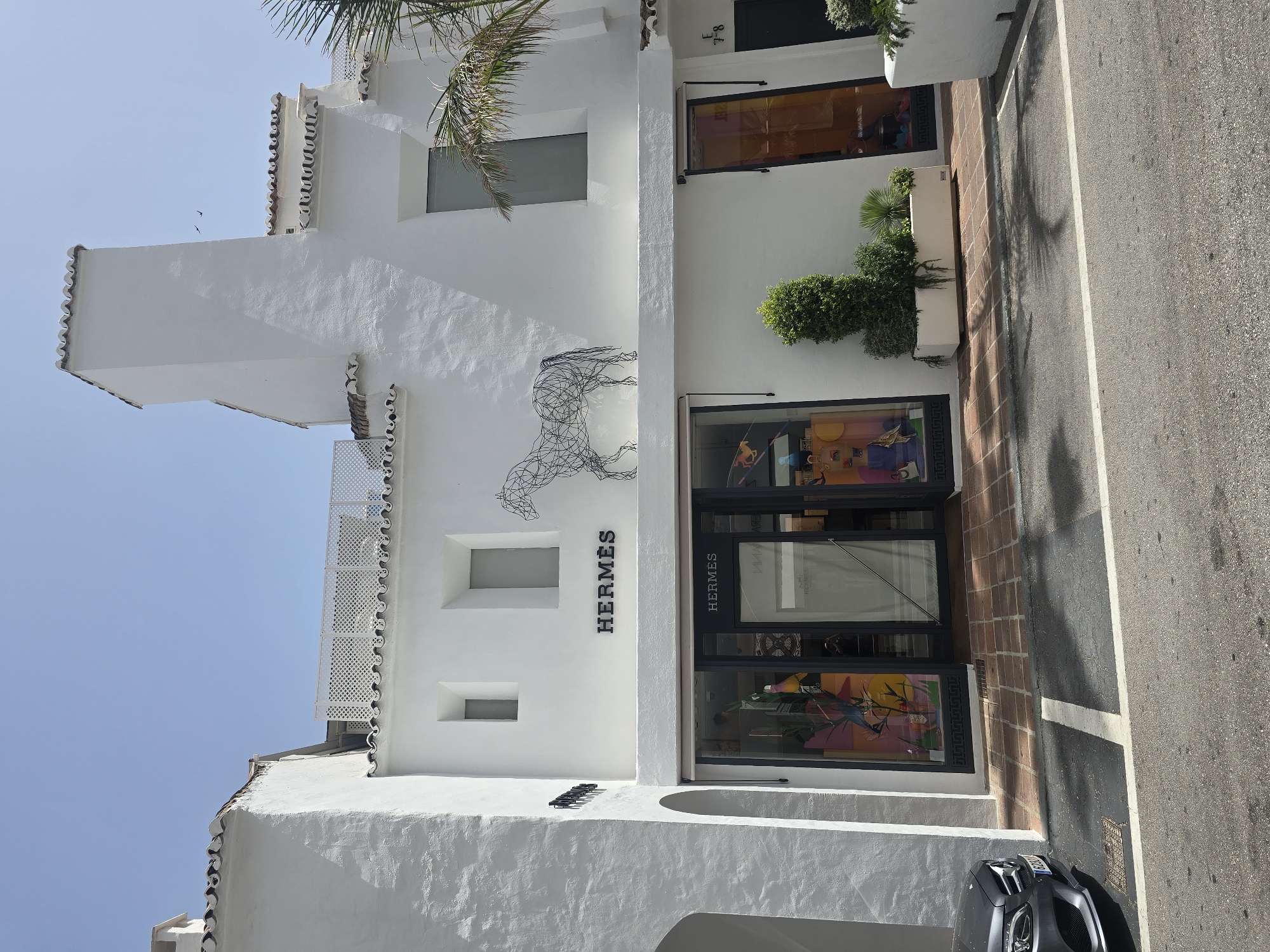 The image shows the exterior of an Hermes store in Marbella, Spain. The building is white with traditional Spanish tile roofing. Above the store entrance, a wire sculpture of a horse is mounted on the wall. The store's windows display colorful merchandise, and a potted plant arrangement sits in front of the entrance. A grey Mercedes-Benz is parked to the left of the building. The street in front of the store is paved, and palm tree shadows are visible on the ground. The sky is clear and blue.