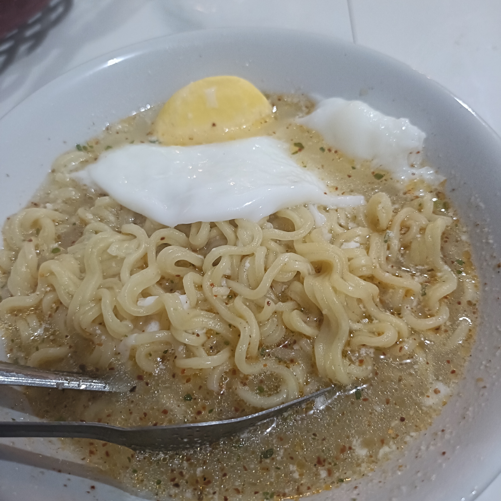 A white bowl filled with noodles in broth and two eggs, one with a hardened yolk and one soft-boiled, rests on a white surface. A set of metal tongs is visible in the lower left portion of the bowl. The location is likely Cangkring, Indonesia.