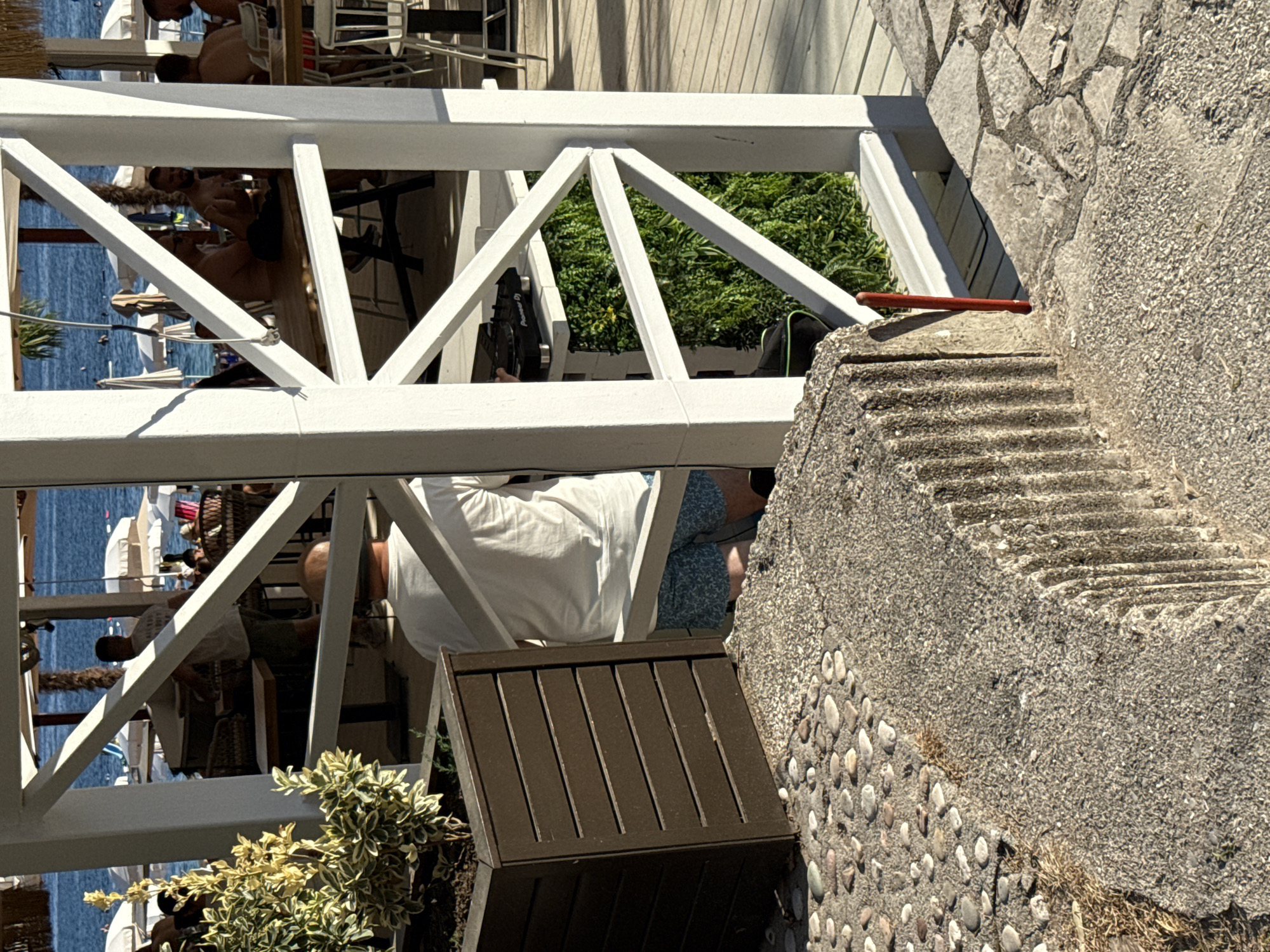In Kamenovo, Montenegro, a scene unfolds at what appears to be a beachside establishment. The foreground is dominated by textured concrete structures and a path of small, rounded stones. Behind this, a dark wooden planter box holds a variegated plant with yellow and green leaves. A white wooden pergola structure cuts through the midground, offering glimpses of the scene beyond.
Through the pergola, several individuals are visible. One person, seen from the back, wears a white t-shirt and patterned shorts. To their right, another person is seated at a table, their face obscured. Further into the background, more people are seated at tables, some appearing to be dining or socializing. Beach umbrellas and the blue sea are visible in the distance.
On a ledge within the pergola, a DJ mixer, specifically a Pioneer DJ device, is set up. A vertical green wall, possibly artificial foliage, serves as a backdrop behind the DJ equipment. The overall impression is one of a relaxed, outdoor setting, likely a restaurant or bar, situated near a beach. The lighting suggests it is daytime, with sunlight casting shadows.