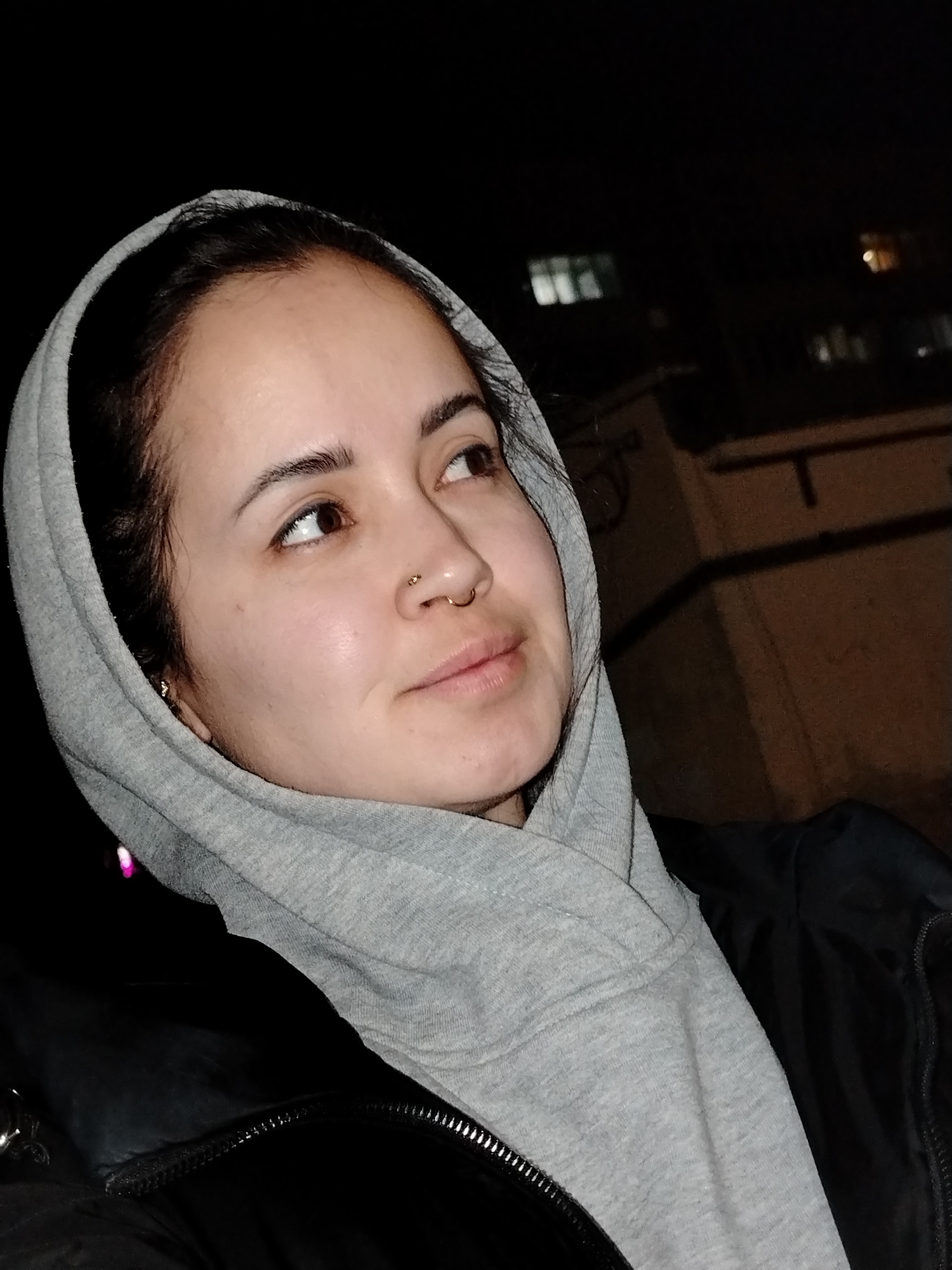 A medium-angle shot shows a person with light skin, brown eyes, and dark hair framed by a gray hooded sweatshirt, wearing a black jacket with a visible zipper. The person has a gold nose ring and a small gold earring. The background suggests an urban nighttime setting, with illuminated windows of a building visible above and to the right of the person's head. The location is Noviy, Russia.
