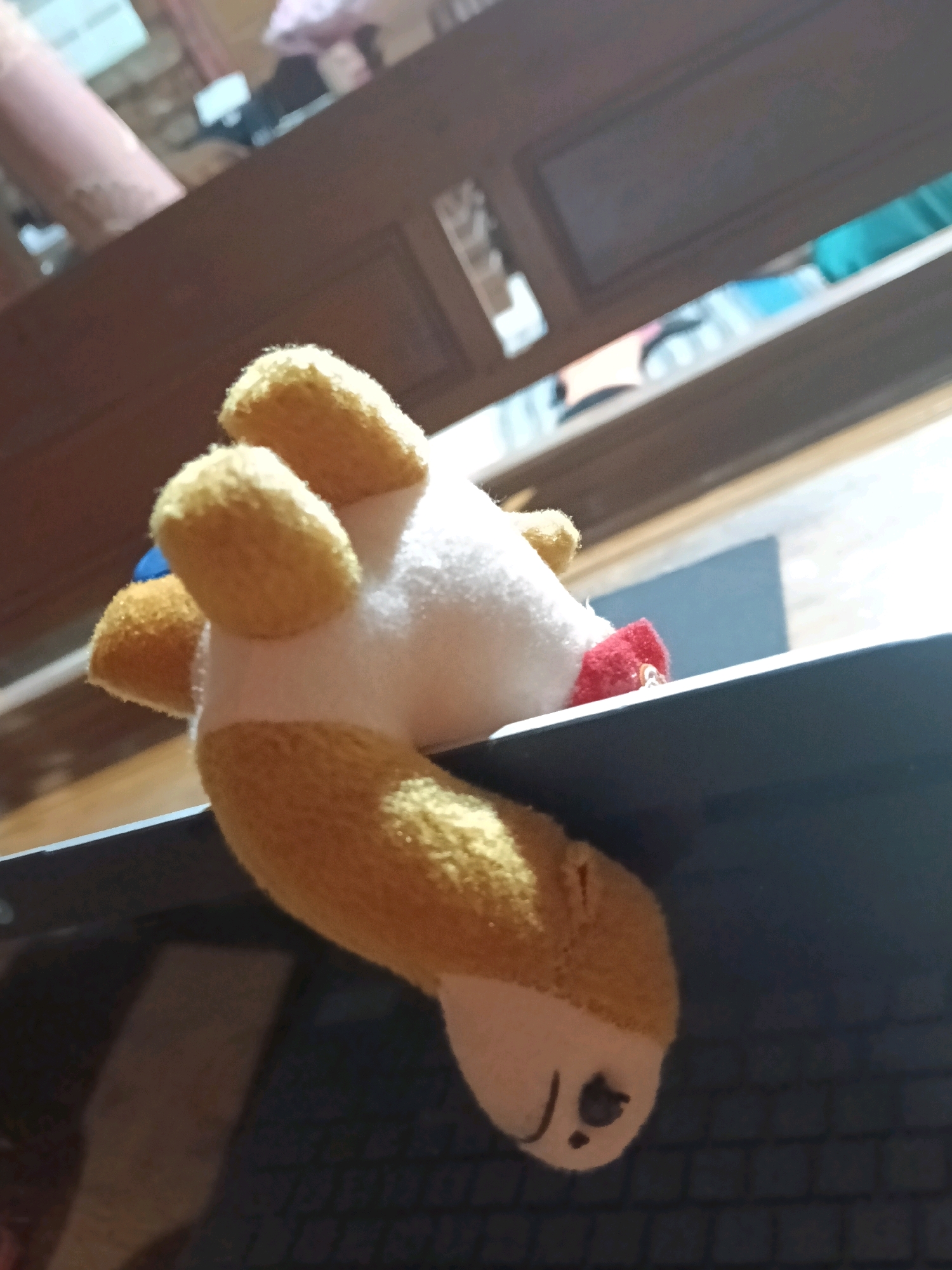 A close-up, low-angle shot focuses on a stuffed toy dog hanging over the edge of a laptop. The toy is predominantly white with tan-colored ears and feet. A small red piece of fabric is visible near its neck. The laptop is black, and its keyboard is partially visible. The background suggests an indoor environment with wooden furniture and some blurred objects, including what appears to be a pink cylindrical object and a shelf with various items. The scene is lit from above, casting shadows and highlighting the texture of the toy. The image does not provide any specific details about a location in the city of Galuk, Indonesia.