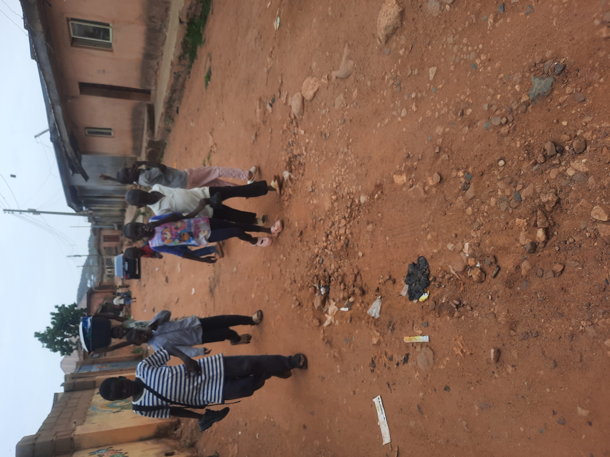 The scene is a dusty, unpaved street in what appears to be a residential area. A group of children are walking along the street. The buildings are simple, likely residential structures. The ground is mostly red dirt and stones with some trash scattered around. The location is Jos, Nigeria.