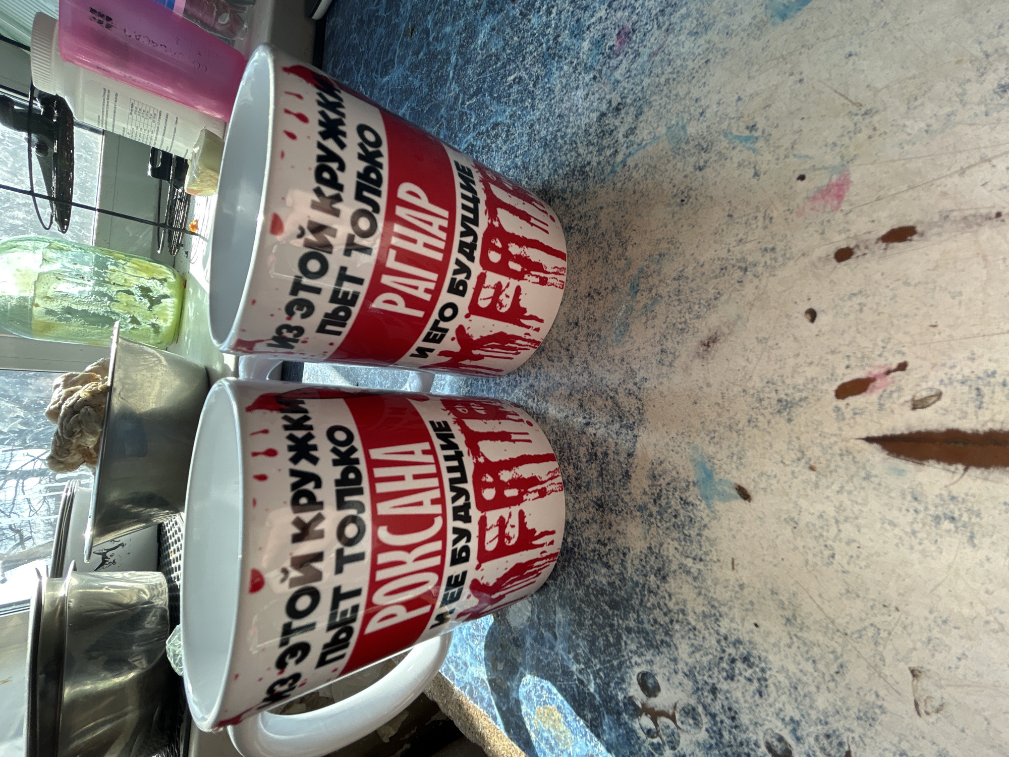This image shows two white ceramic mugs with a red band printed with Russian text. The text appears to indicate that the mug is specifically for a person named "Roxana" and another for "Ragnar" respectively, and that only they can drink from it. The mugs are sitting on a patterned surface with a dark blue and white color scheme. They appear to be positioned near a window, which is visible in the background along with other items, including a green jar, a pink container, and some metallic cookware. The scene seems to be inside a kitchen. The location of the scene is not determined.