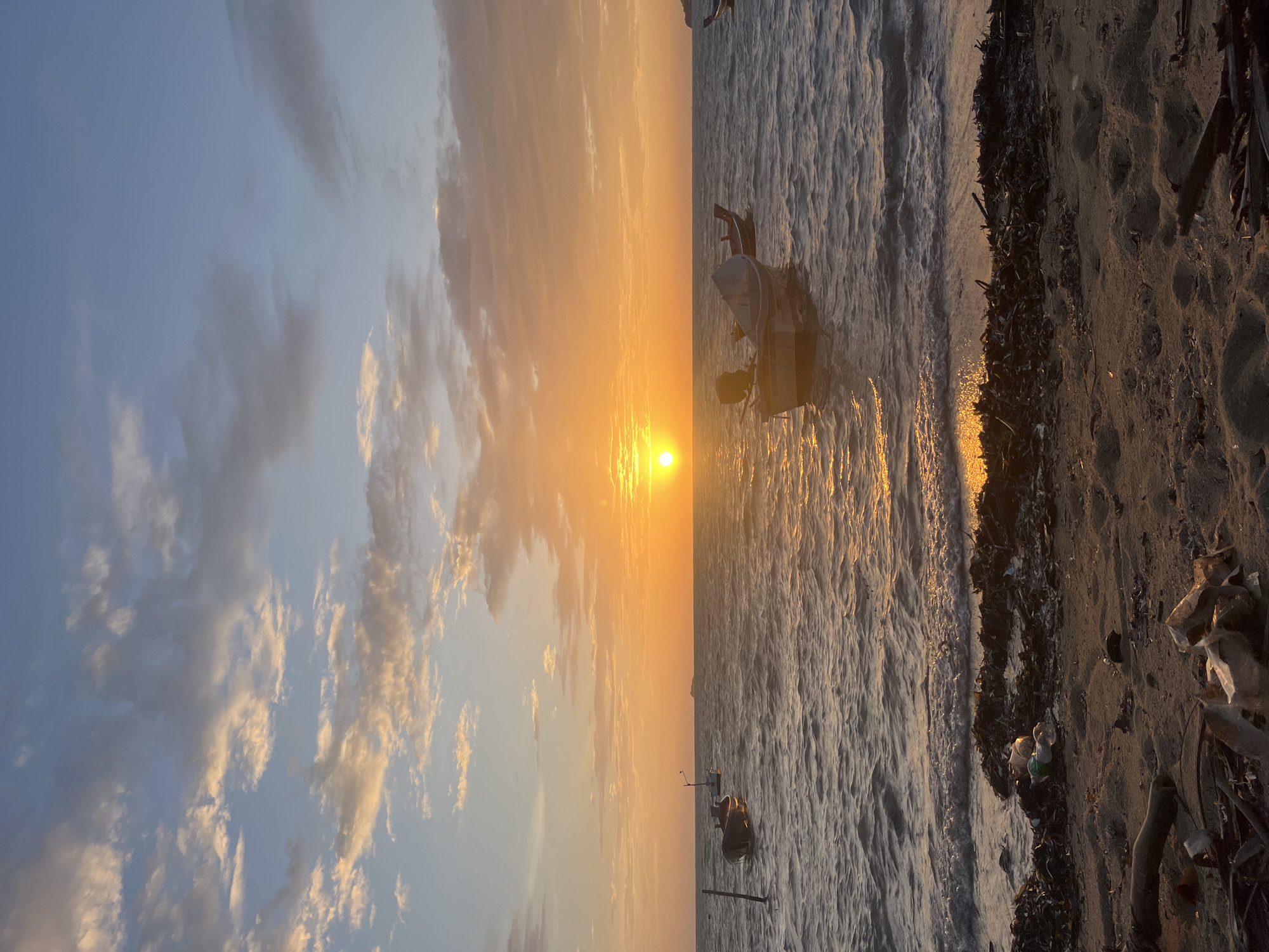 The image depicts a beach scene at sunset in Ko Pha-ngan District, Thailand. The sun, a bright orb of orange, is low on the horizon, casting a warm glow across the sky and water. Clouds, tinged with orange and pink, are scattered across a pale blue sky. The sea is choppy with small waves, reflecting the sunset's colors. Several small boats are anchored offshore, bobbing gently in the water. The foreground is a sandy beach littered with debris, including plastic bottles, wood fragments, and seaweed. The overall atmosphere is serene, yet the presence of litter suggests environmental concerns.
