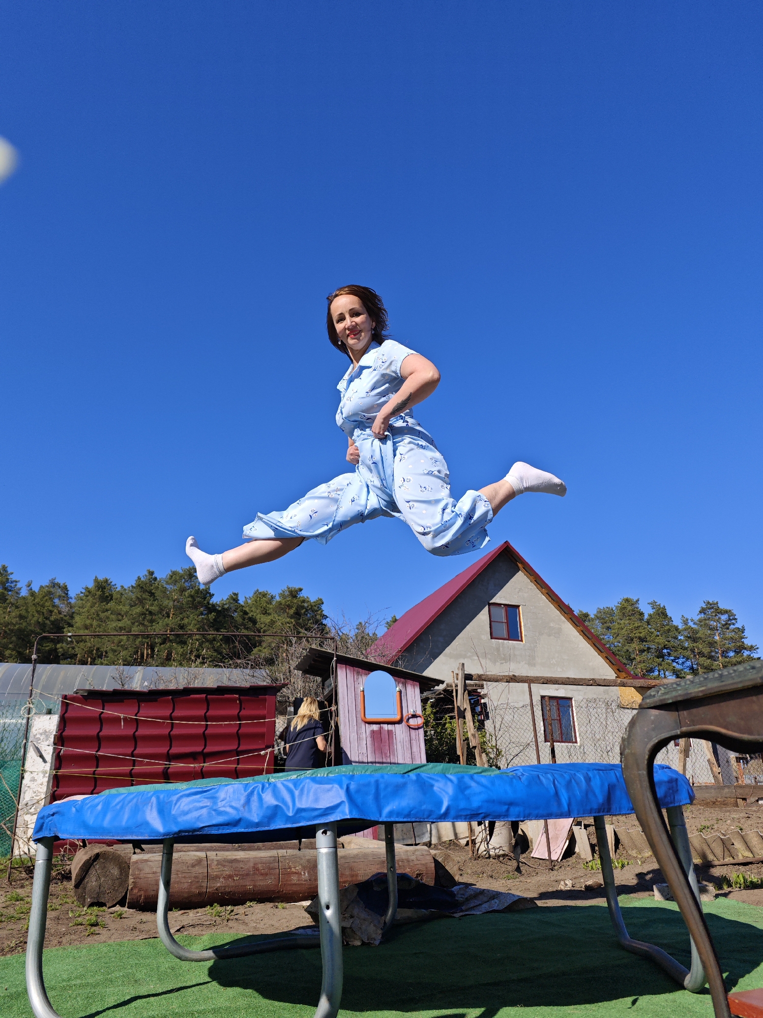 A woman is captured mid-air, seemingly jumping on a trampoline outdoors in Noviy, Russia. She is wearing a light blue patterned dress and white socks, with her arms slightly bent. The background includes a house with a red roof, trees, a trampoline, and a fence. The sky is clear and blue. A second woman is to the left of the trampoline, standing next to a red structure.