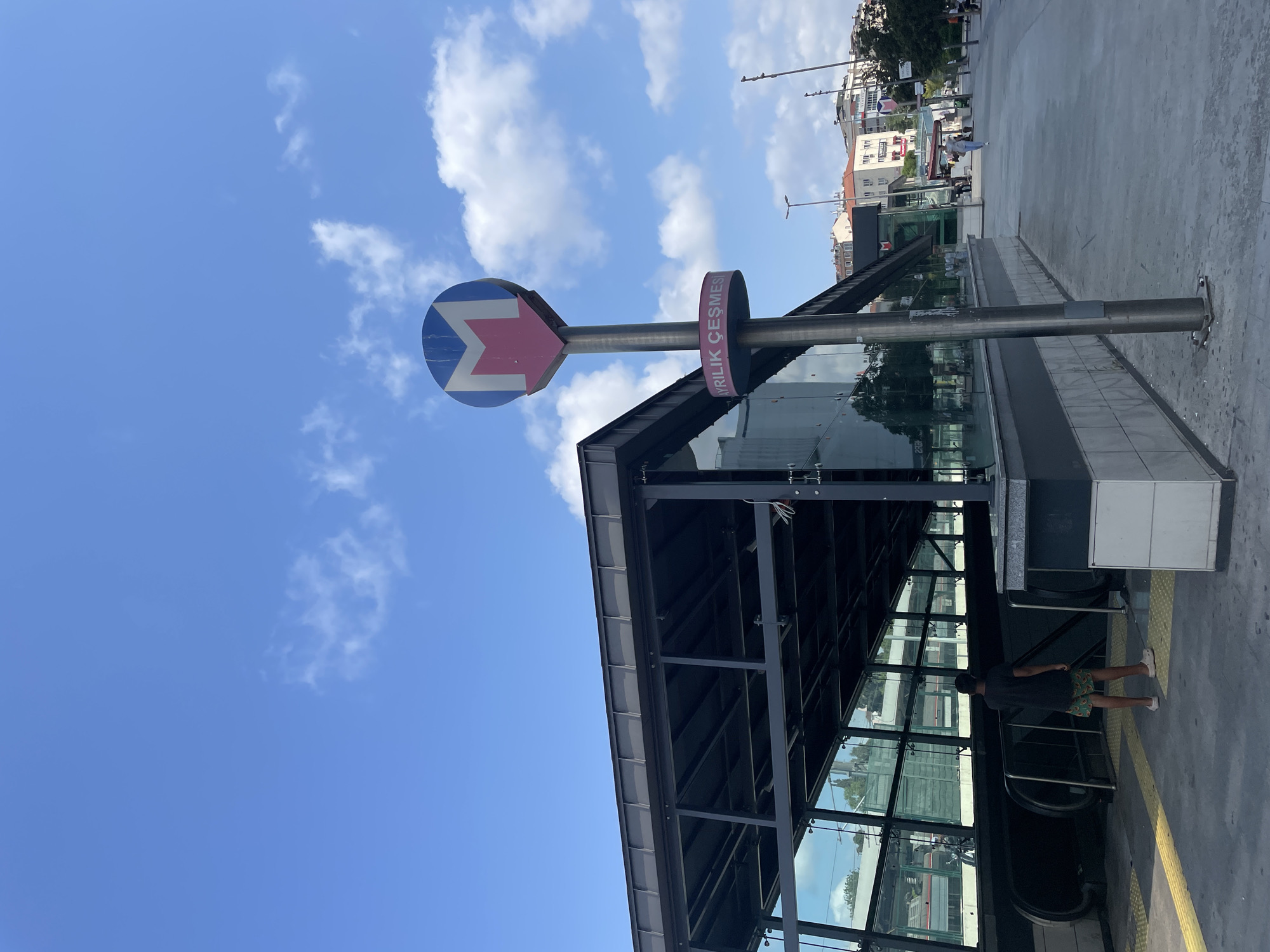 The image shows a street scene in Kadıköy, Turkey, with a metro station entrance prominently featured. A blue and pink metro sign with the letter "M" is visible on a pole, along with text that reads "AYRILIK ÇEŞMESİ". The metro station itself has a modern design with a dark metal roof and glass walls. An escalator leads down into the station, where a young person wearing a dark t-shirt and patterned shorts stands. The street is paved with concrete and yellow tactile paving lines. In the background, buildings of various heights and architectural styles line the street, and trees are visible. The sky is blue with scattered white clouds, suggesting a clear day. Other people are walking in the distance along the street.
