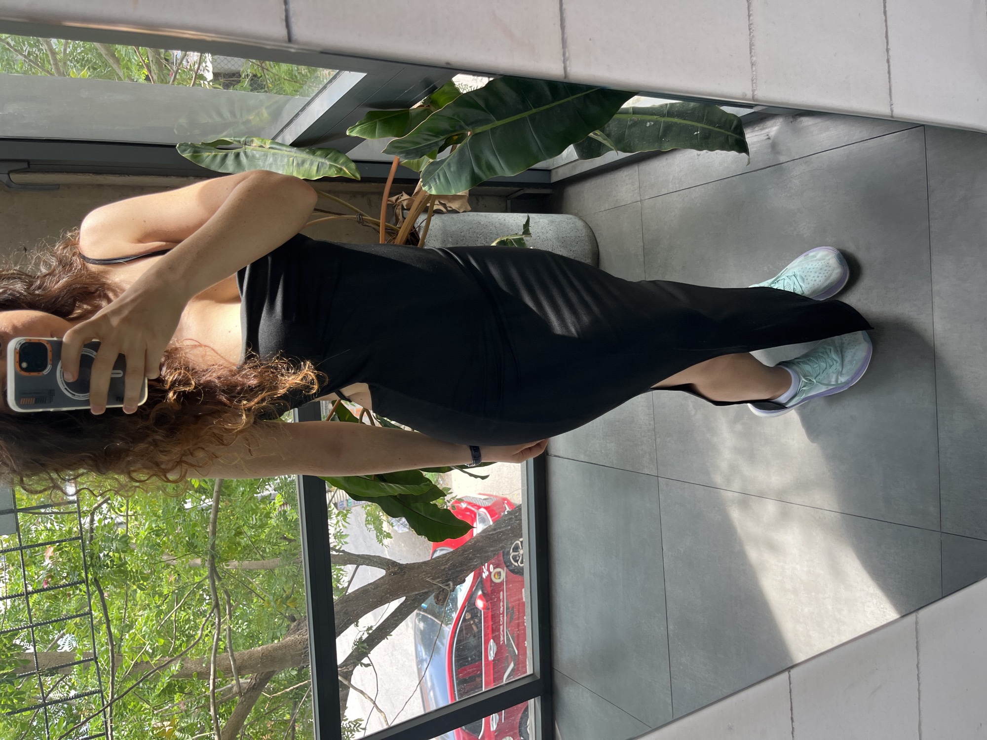 A woman stands in front of a mirror in a residential setting, taking a selfie with her phone. She is wearing a black, form-fitting dress with cutouts on the sides, and light blue athletic shoes. The background shows a window with a view of trees and a red car on a street. A large plant is visible to her right. The floor is tiled with large gray tiles. The lighting suggests it is daytime.